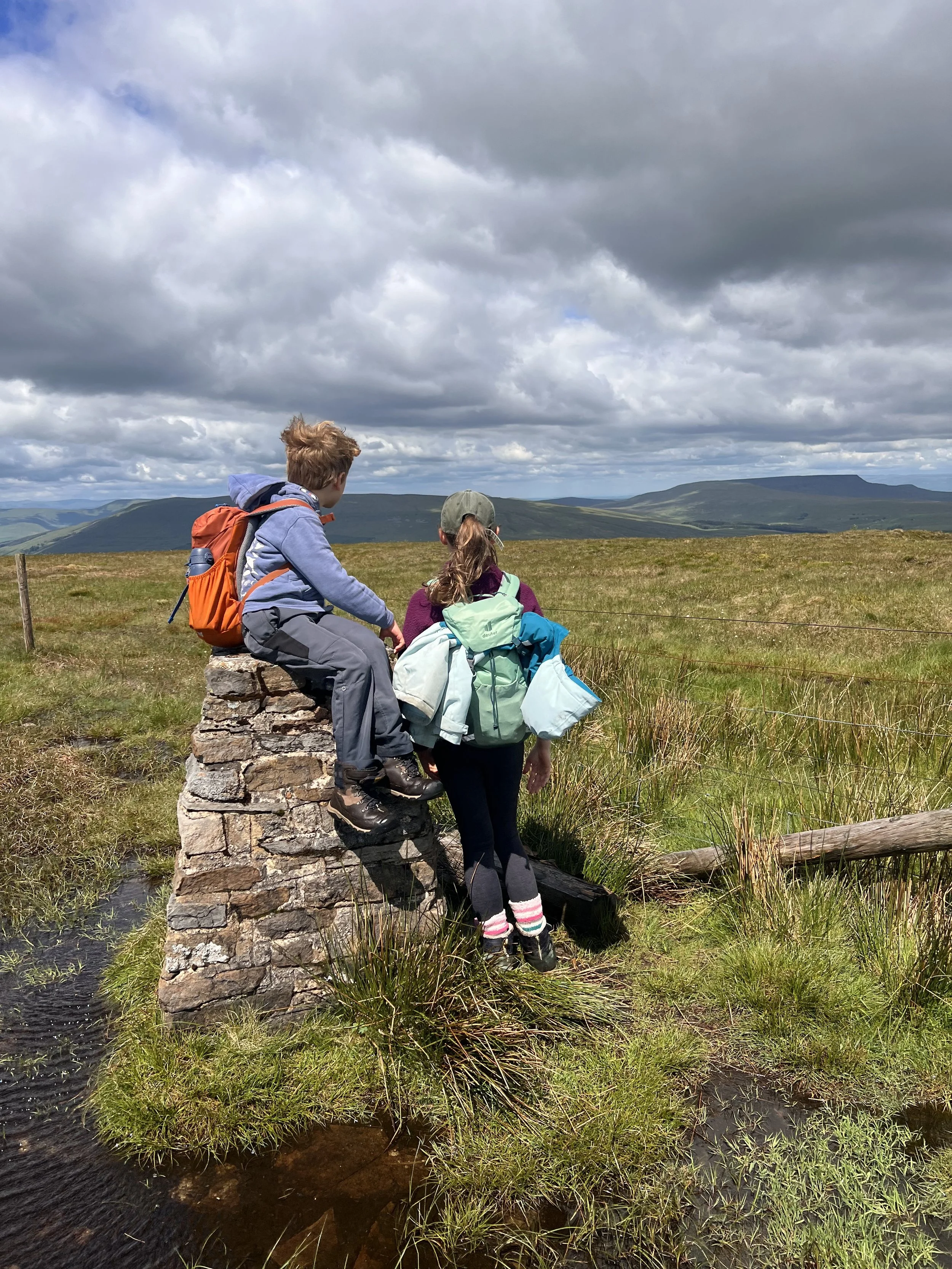Two children sitting on a dry stone wall trig point with mountain views behind