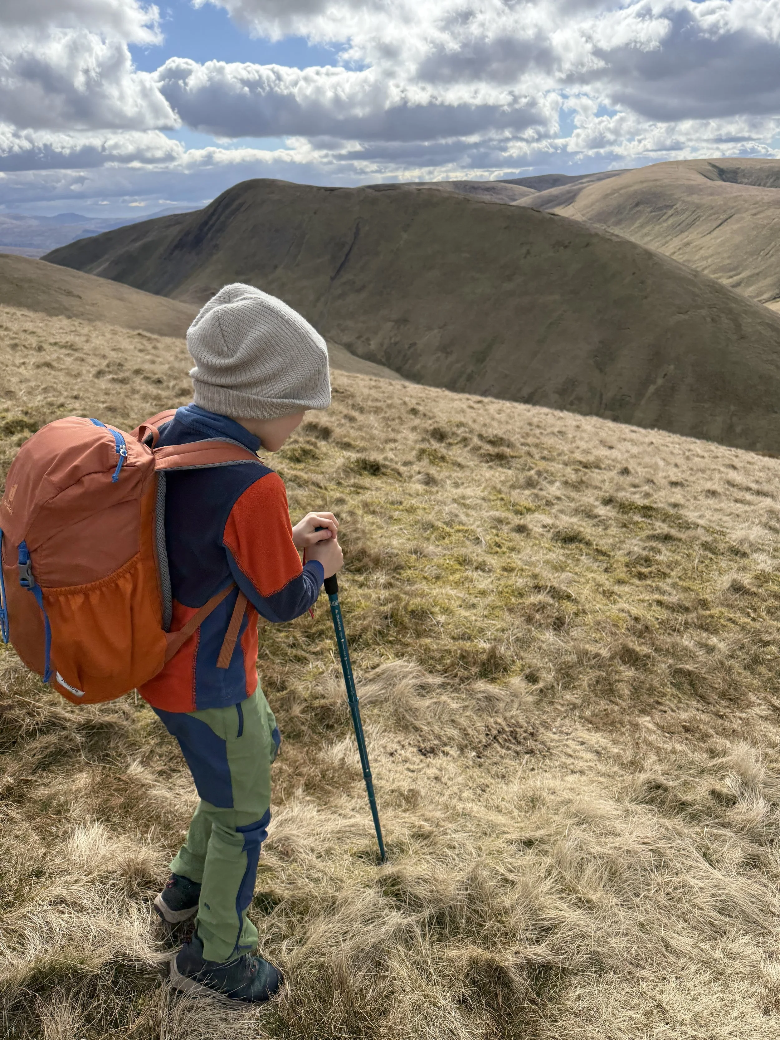 Child standing with rucksack and hiking pole with a beanies hat on his head with views of mountains beyond