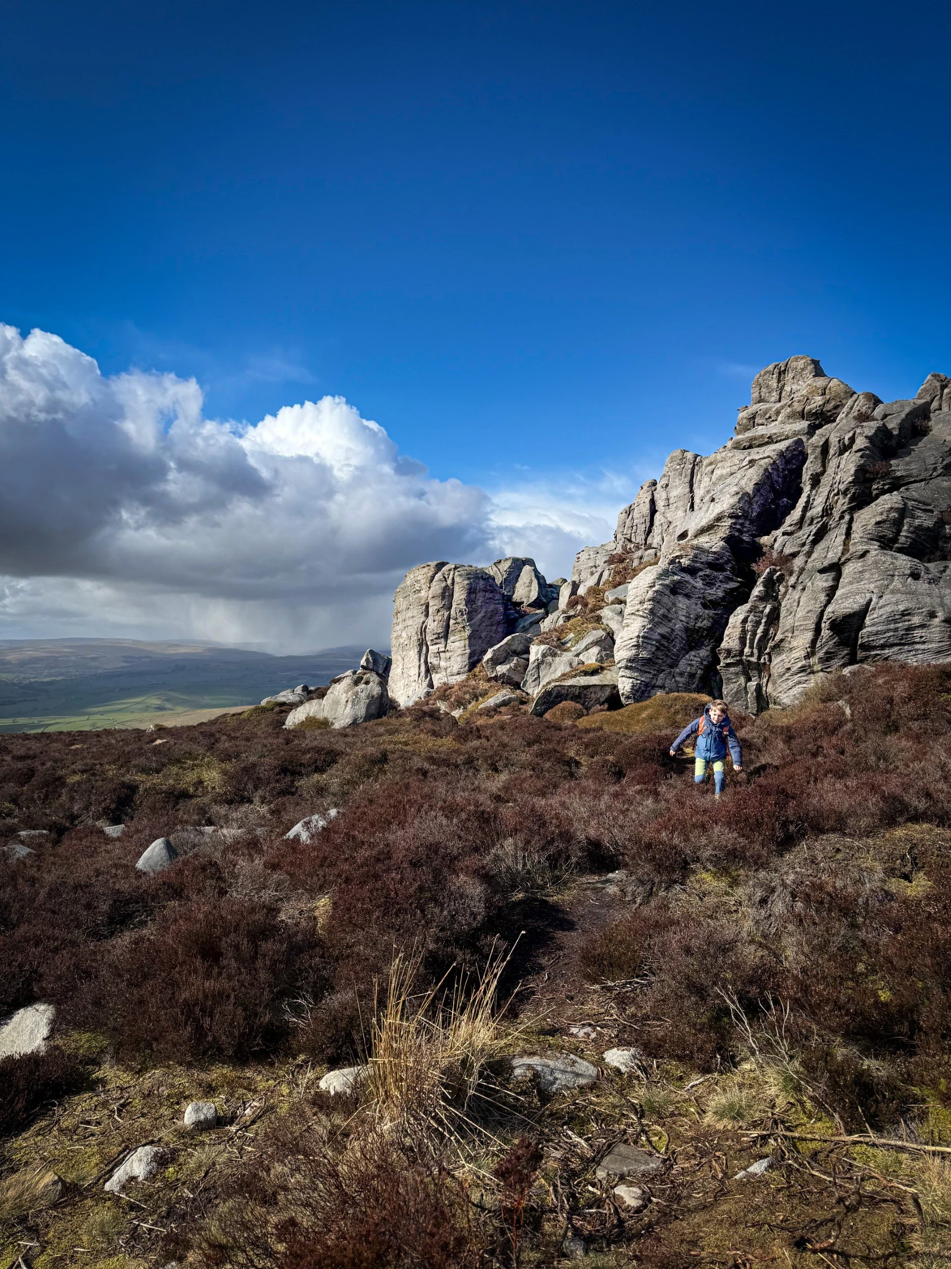 Child walking through brown undergrowth with rocky outcrop behind and blue skies with storm in the distance