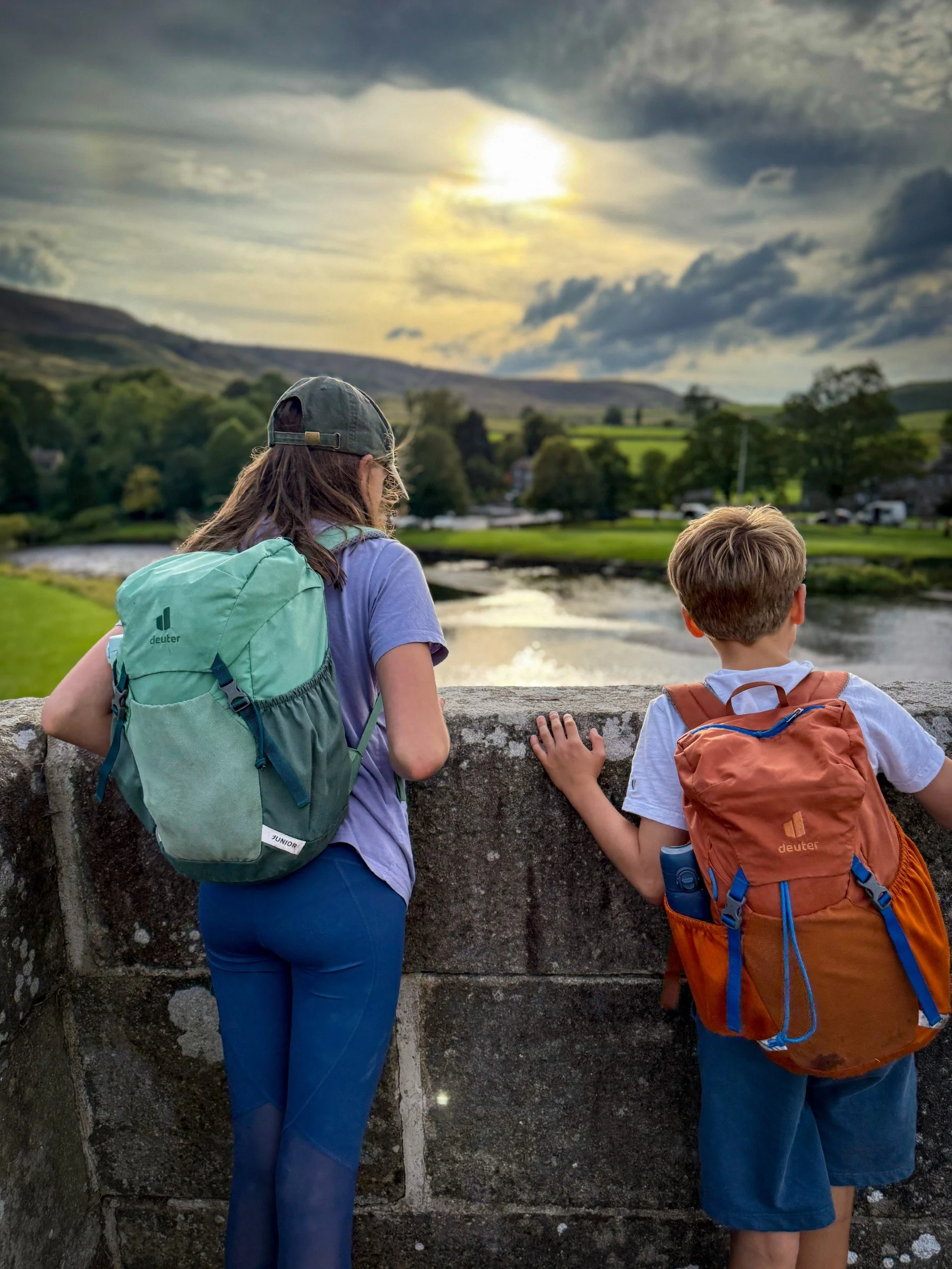 Two children standing on a bridge in summer looking at river with low sun trees and moorland behind