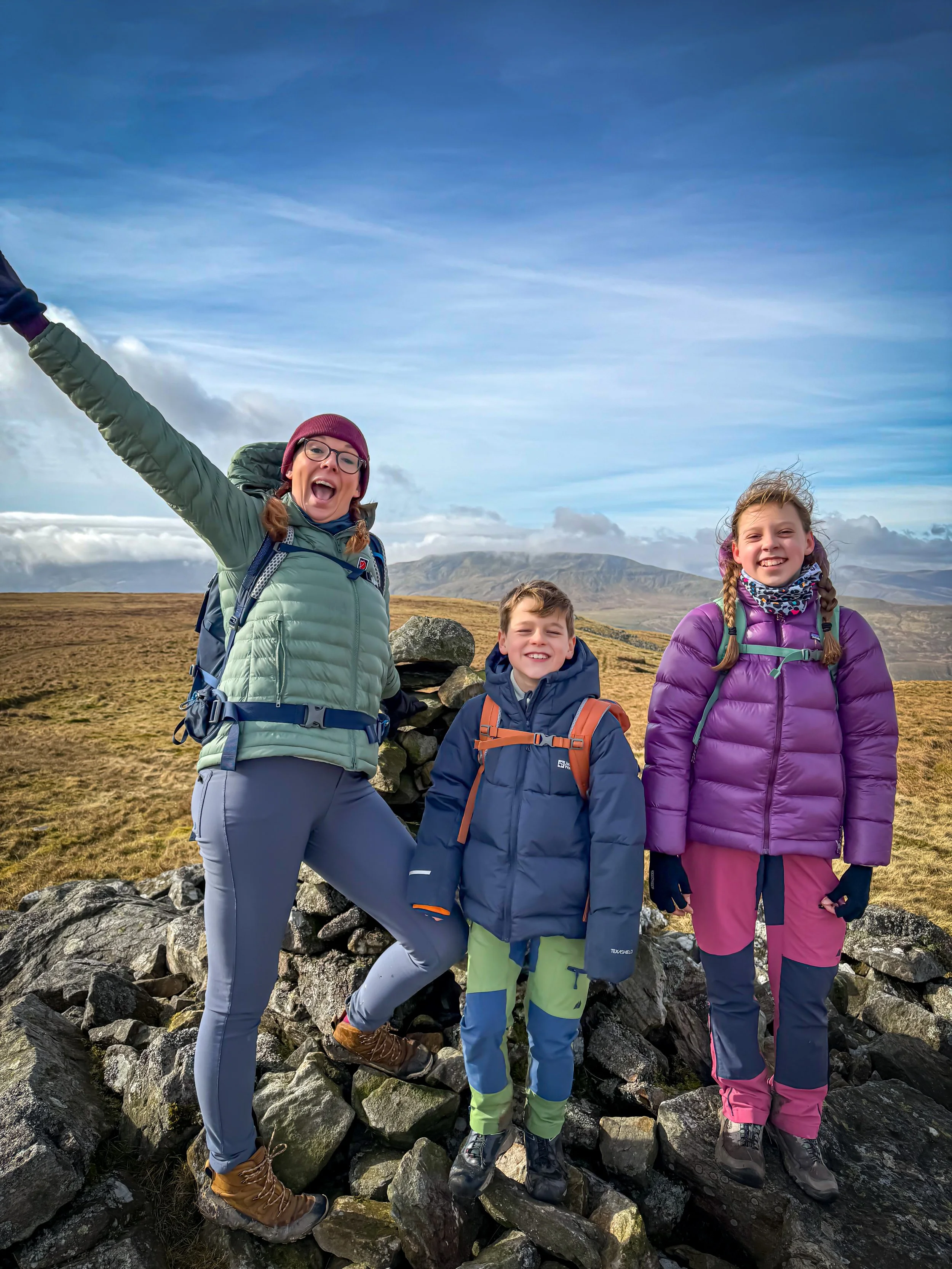 An adult and two children standing next to a stone cairn with mountains behind under blue skies