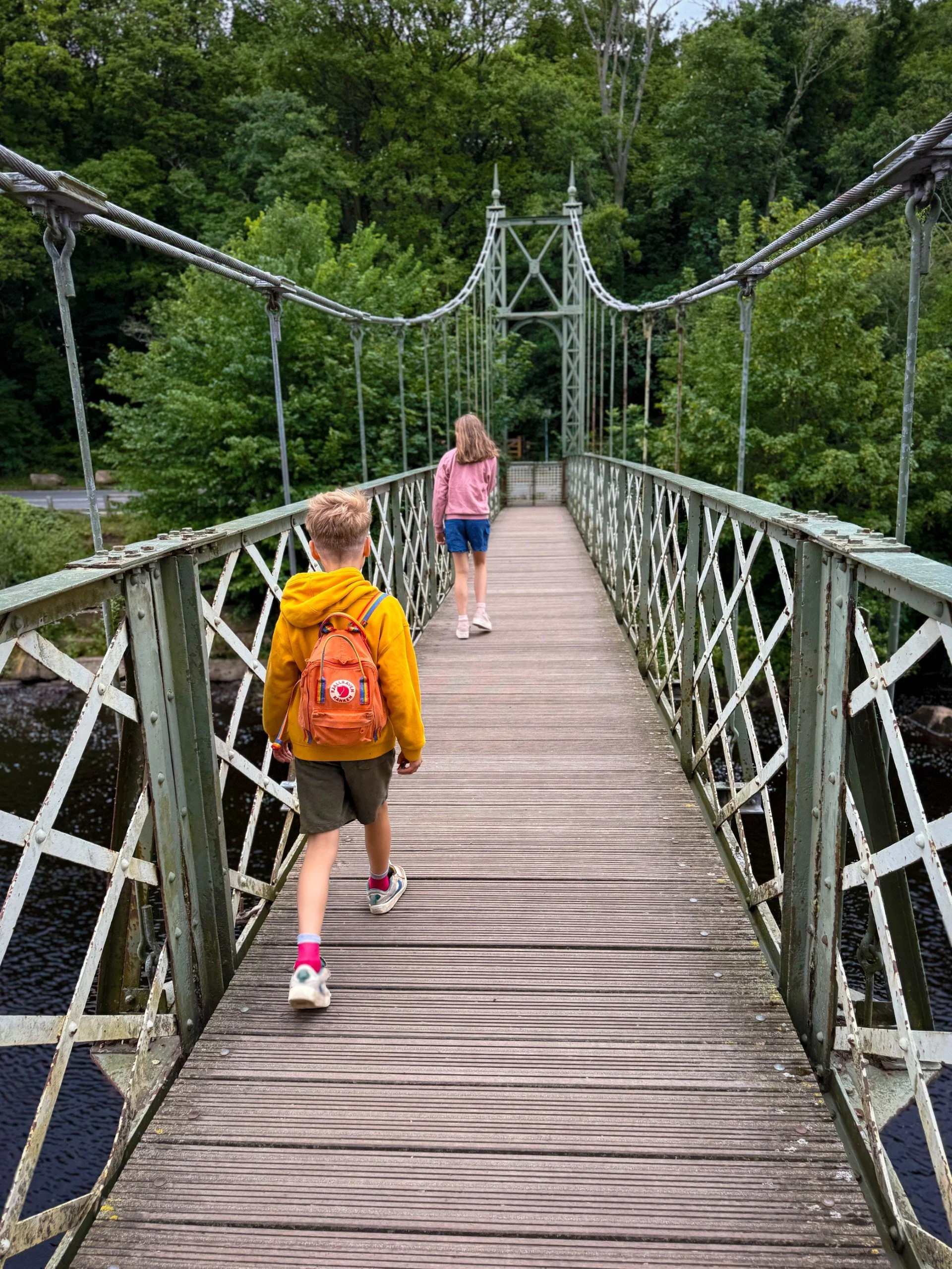 Two children wearing summer clothes walking across an iron suspension bridge with woods behind
