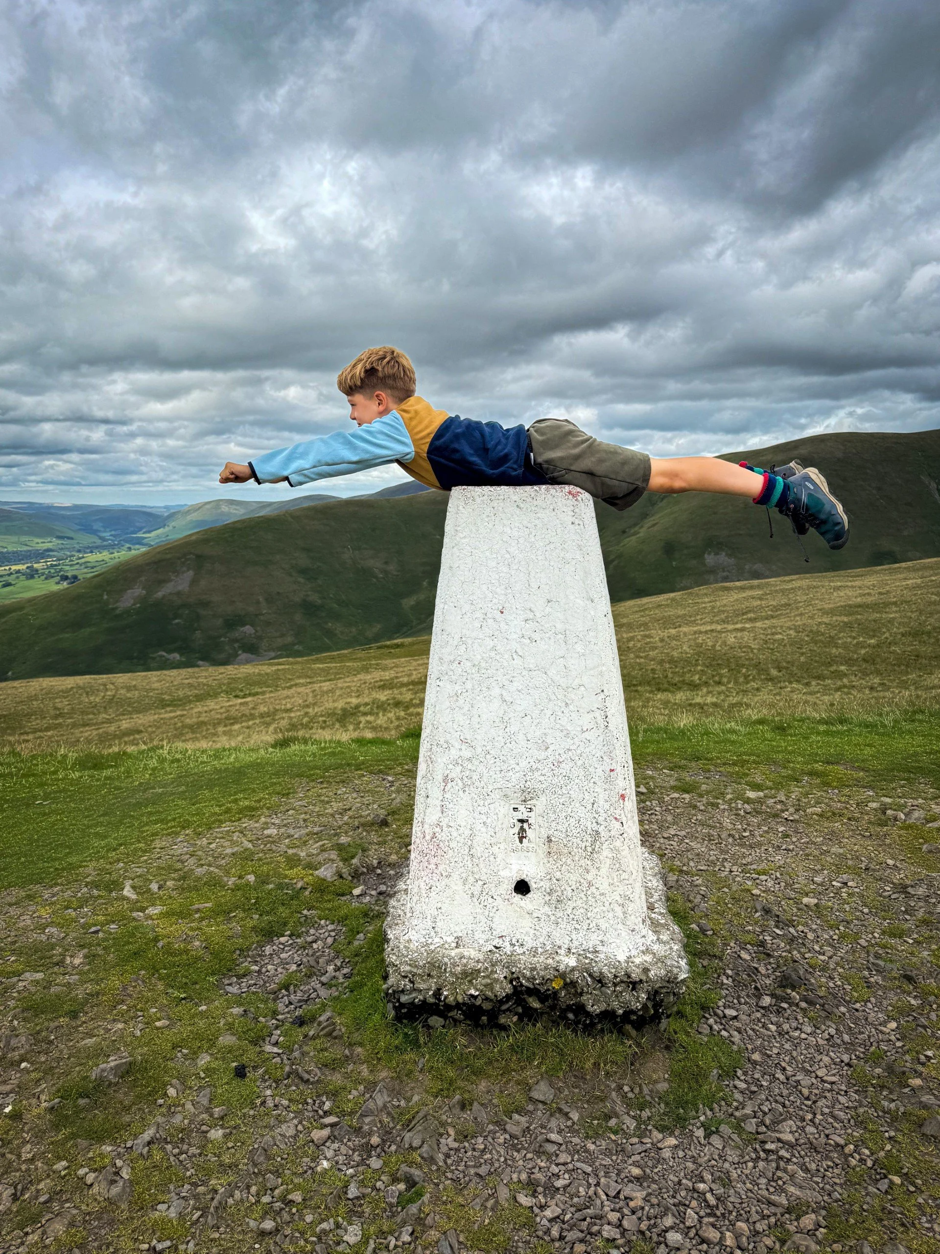 Child laid on top of a trig point in a superman pose with fells behind