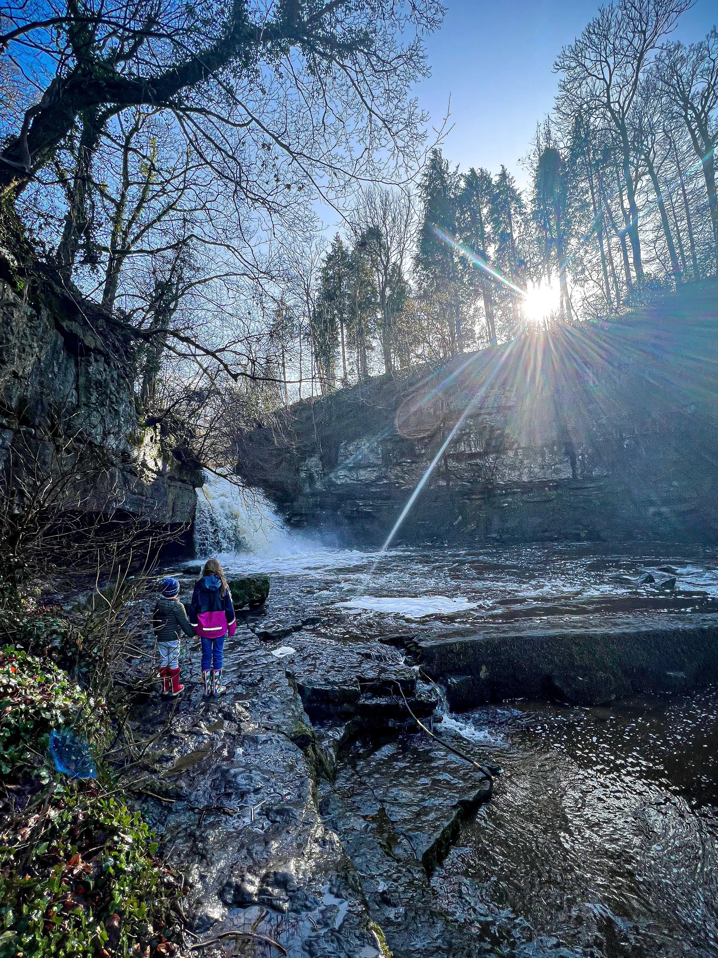 Children standing in front of a raging waterfall with sun peeping through trees