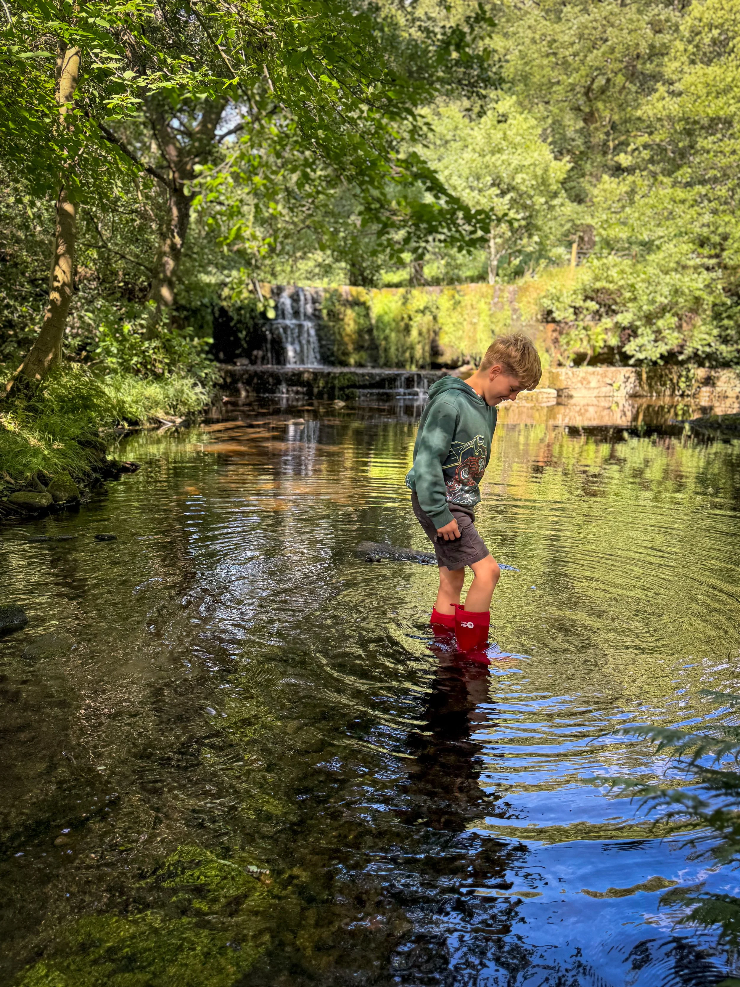 Child in red wellies wading in a river with a waterfall behind and surrounded by trees