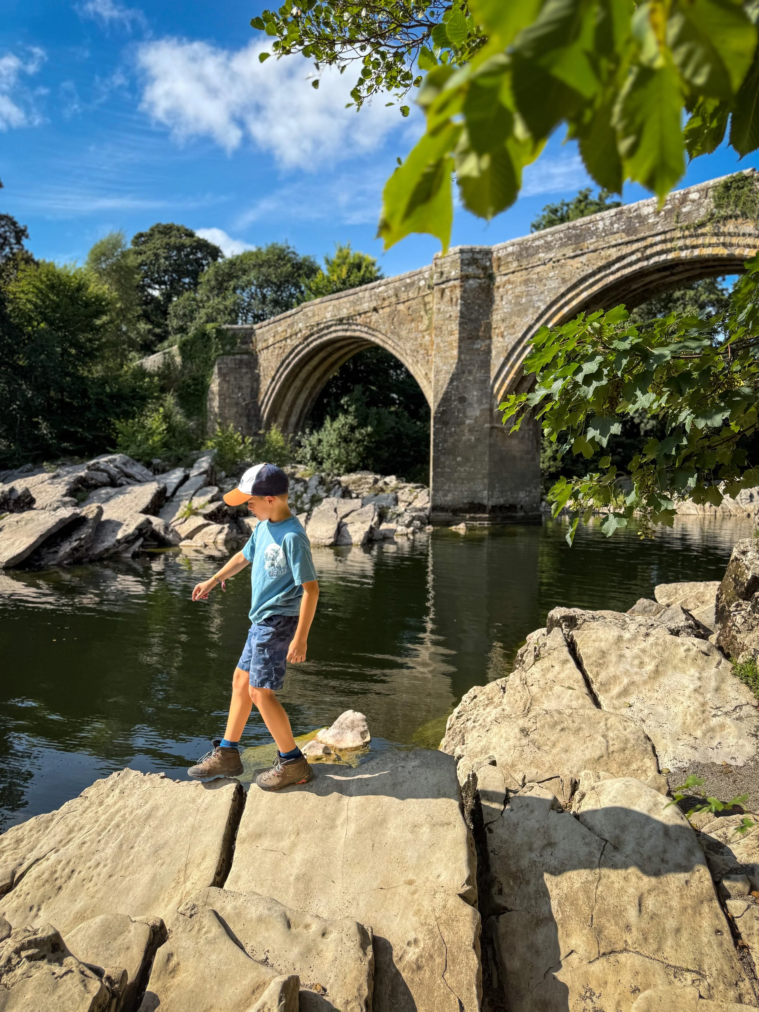 Child walking across a rock under blue skies with trees and a stone bridge