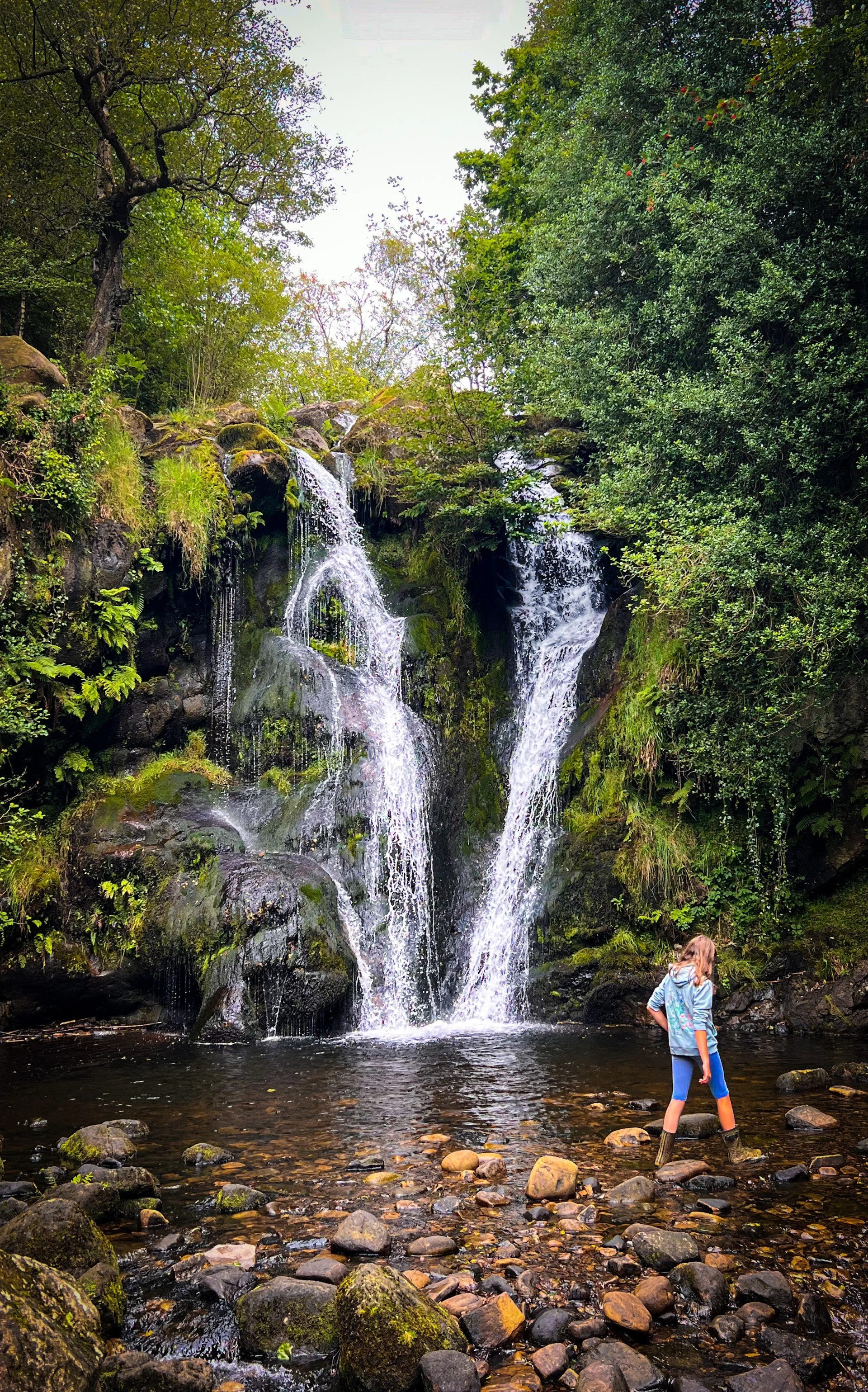 Child walking along the side of a waterfall surrounded by green summer foliage