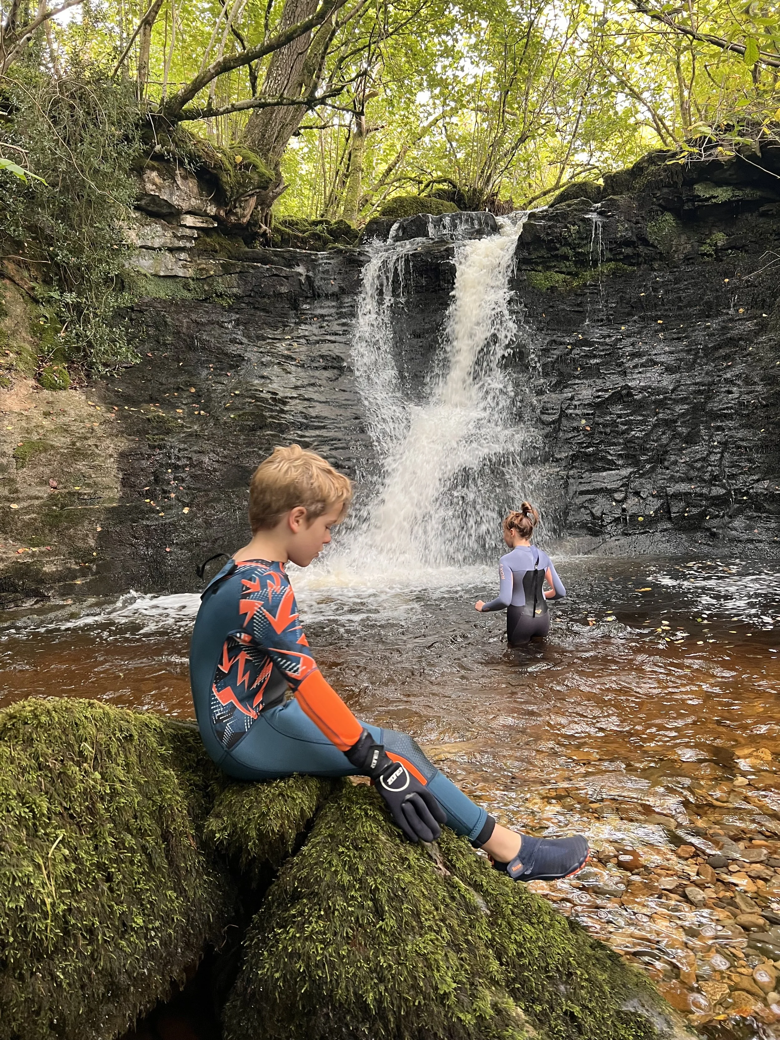 Two children playing in the water of a waterfall one is sat on a mossy rock