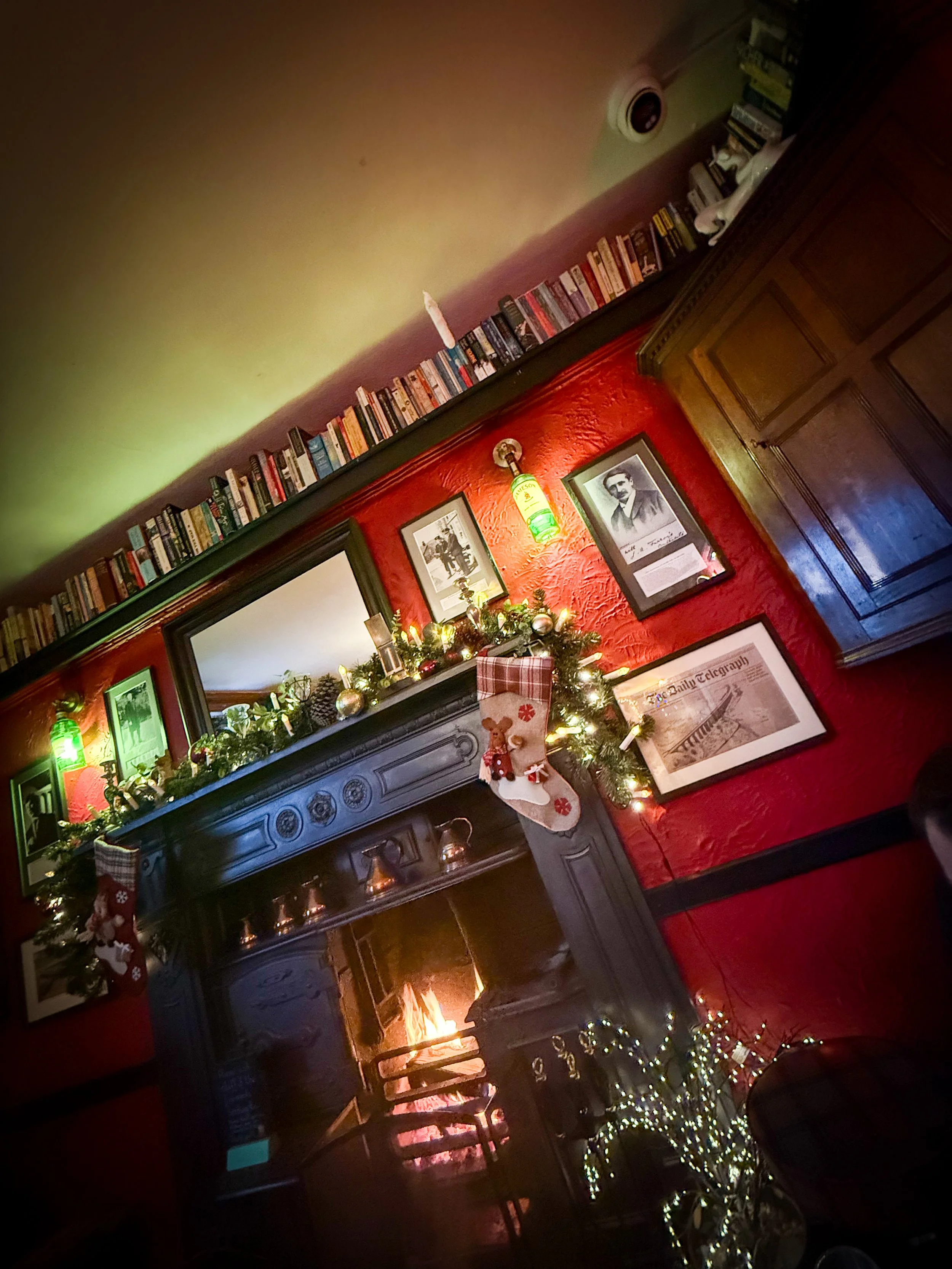 A cosy log fire and books on a shelf with red walls with old fashioned pictures on it in a pub