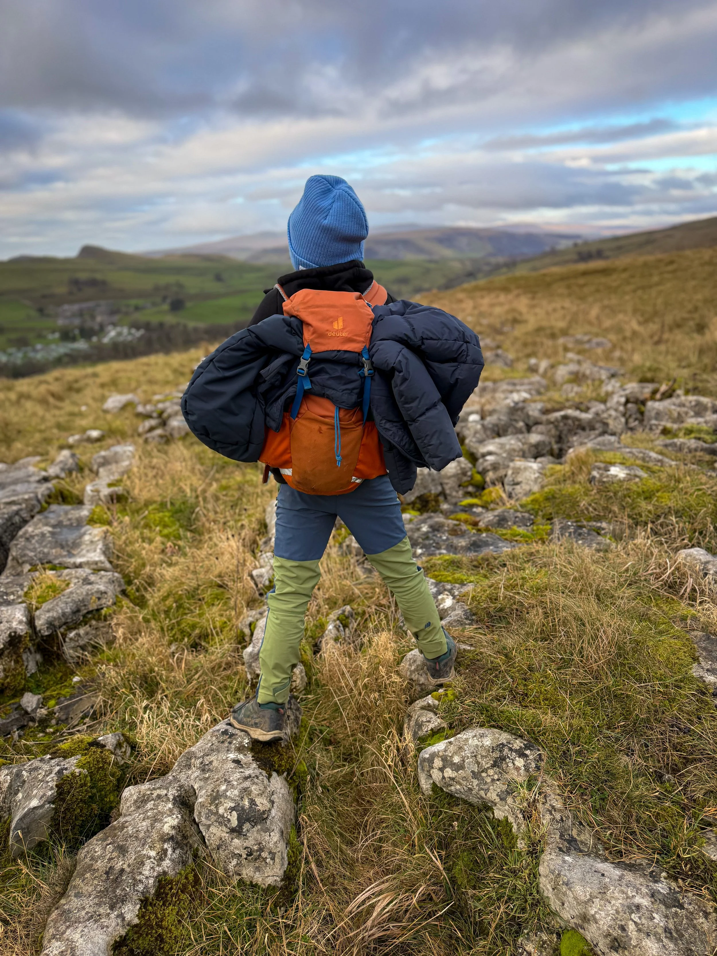 Child with orange rucksack and blue hat standing on rocks looking out to mountain views in winter