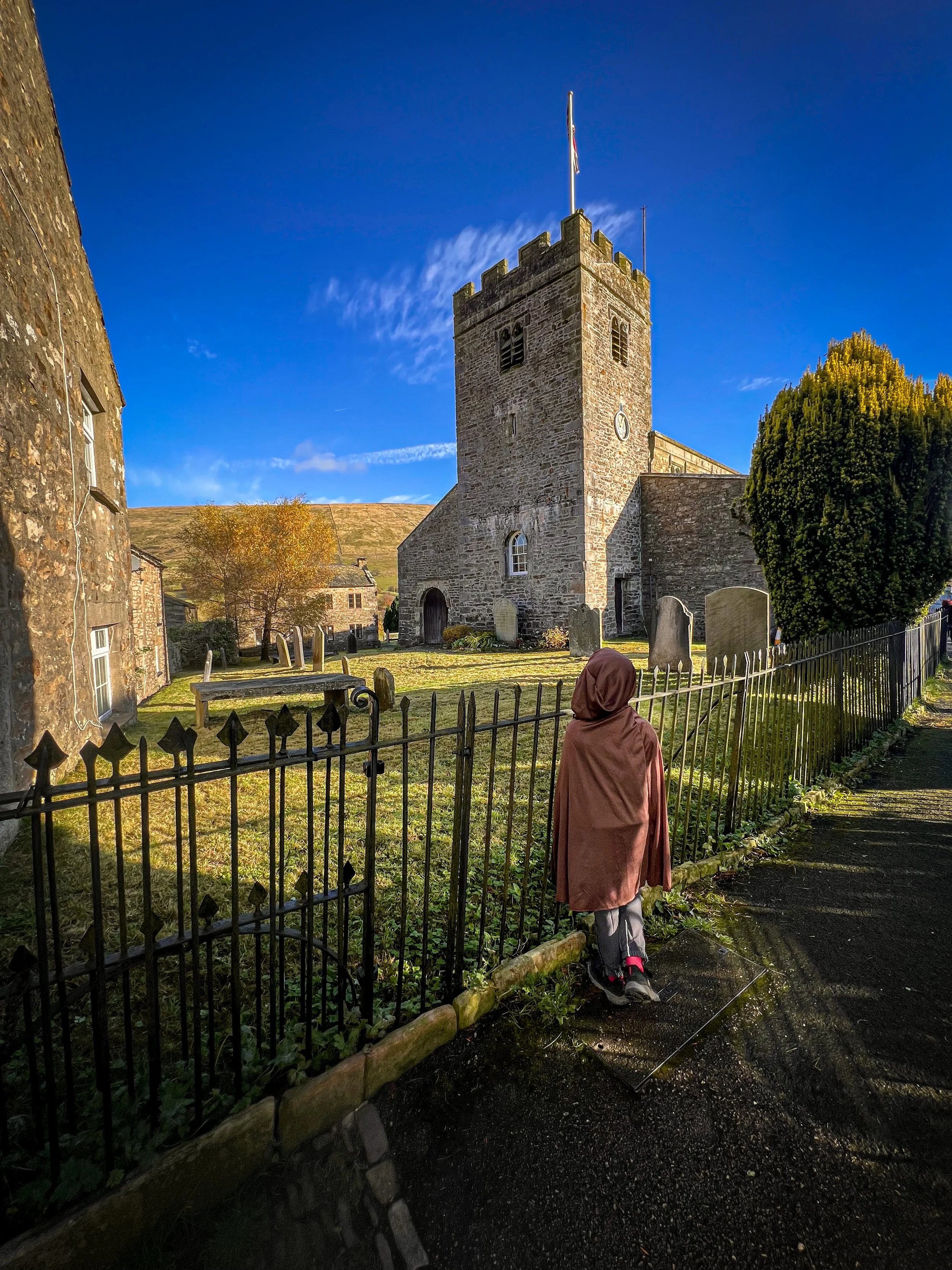 Child standing in a cloak next to a church in a church yard under blue autumn sky
