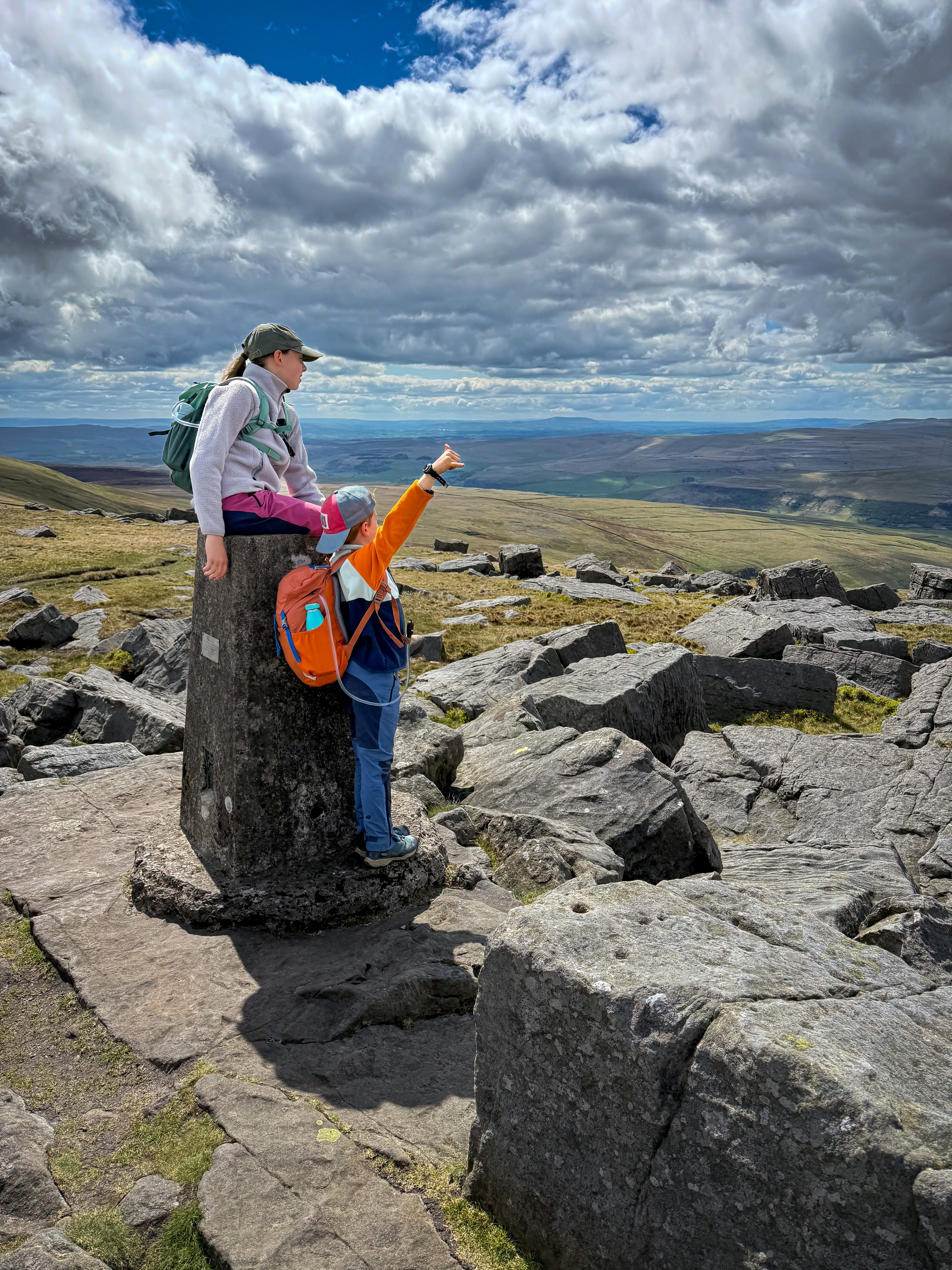 Two children on top of a mountain one with arm in the air the other sat on a trig point loking at view