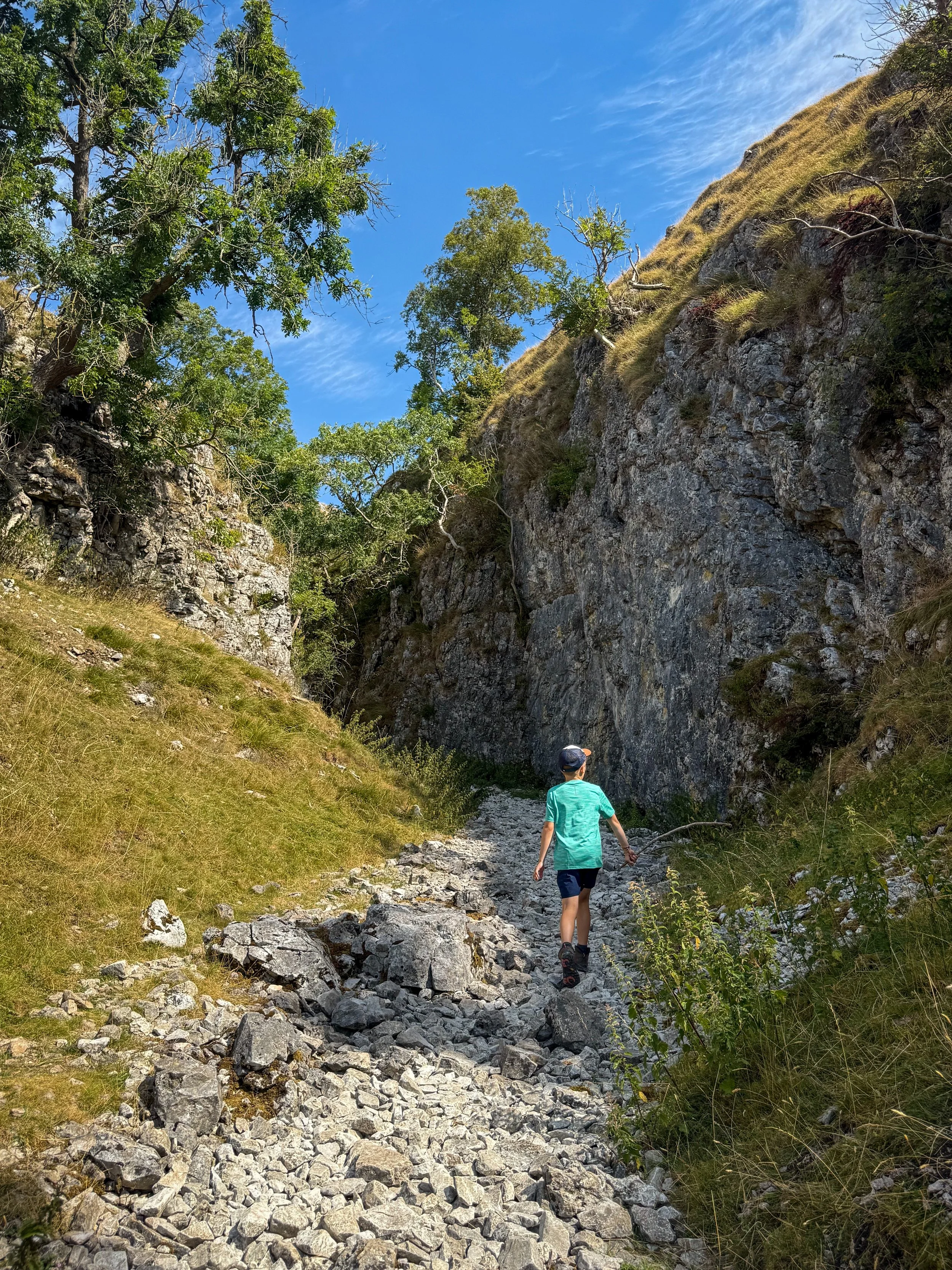 Child in green tshirt walking into a rocky gorge lined by trees and patches of grass under blue sky in summer