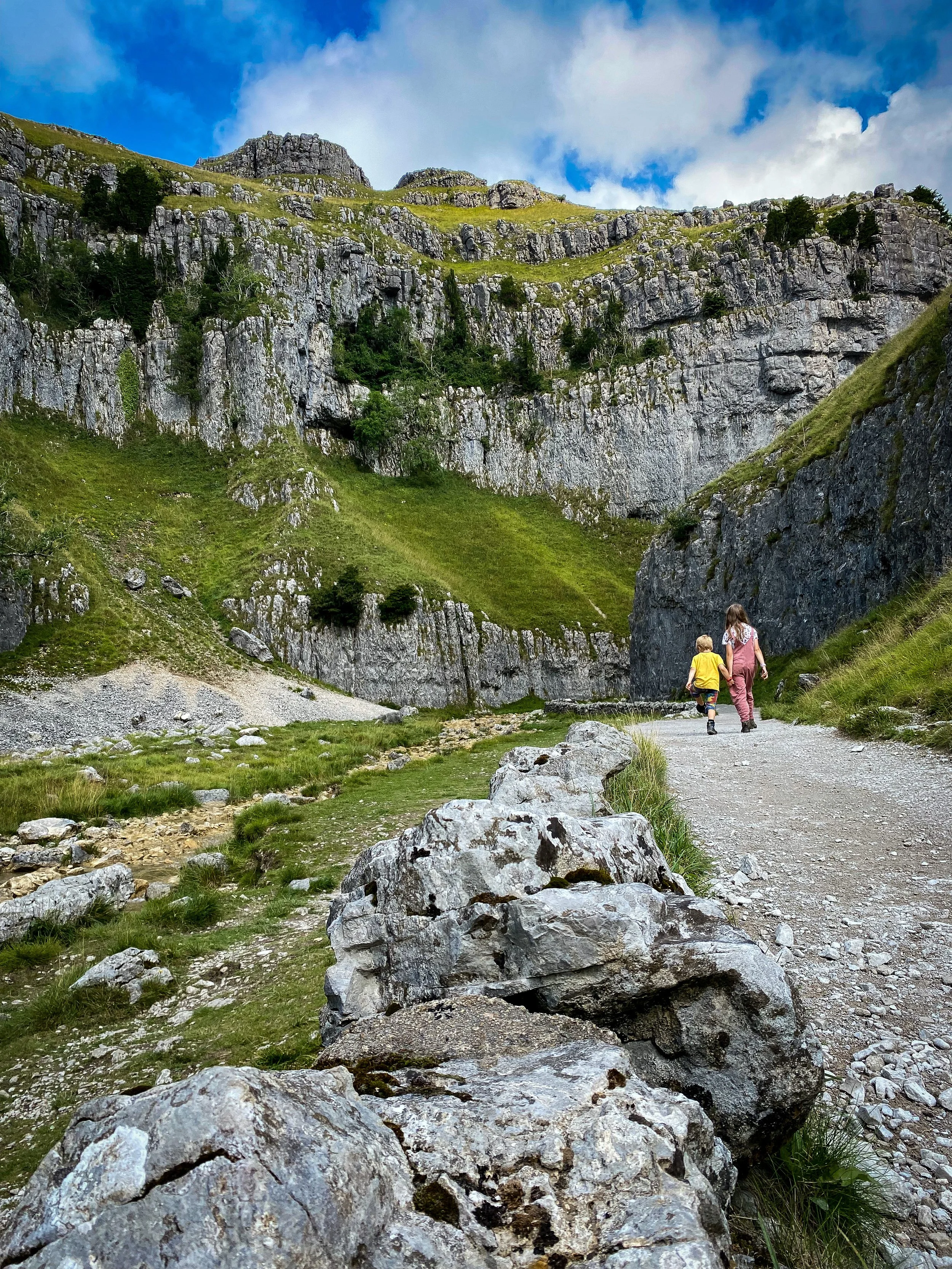 Children walking along stony path towards a large cliff face under blue skies