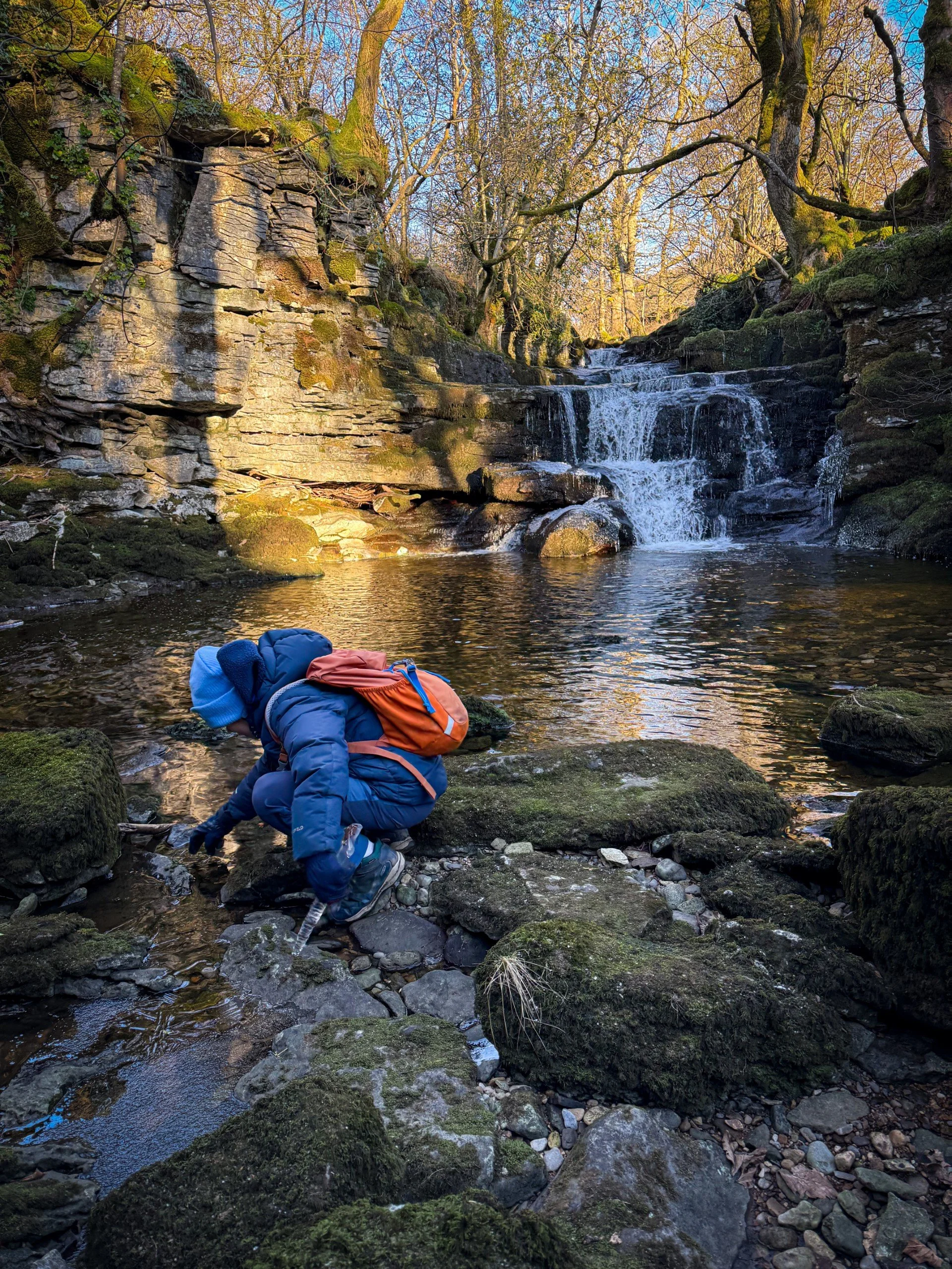 child crouching playing with ice next to a winter waterfall with trees surrounding