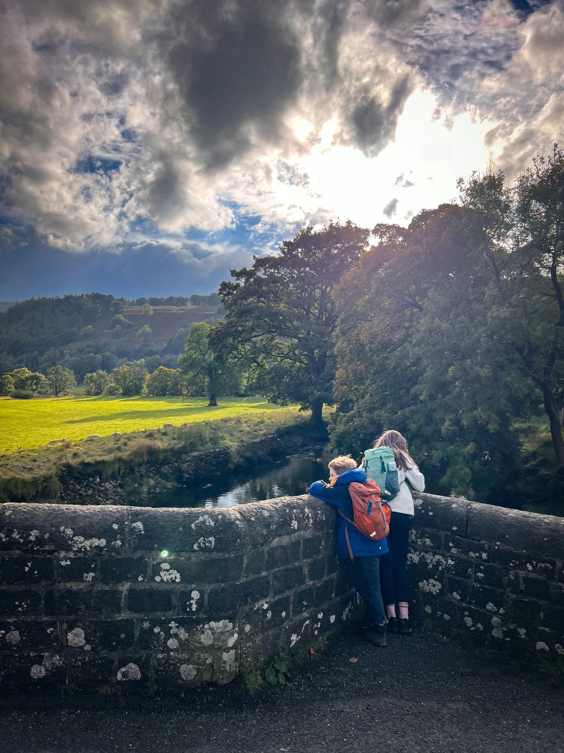 Children standing on an stone built bridge with sun peeping through clouds and late summer trees