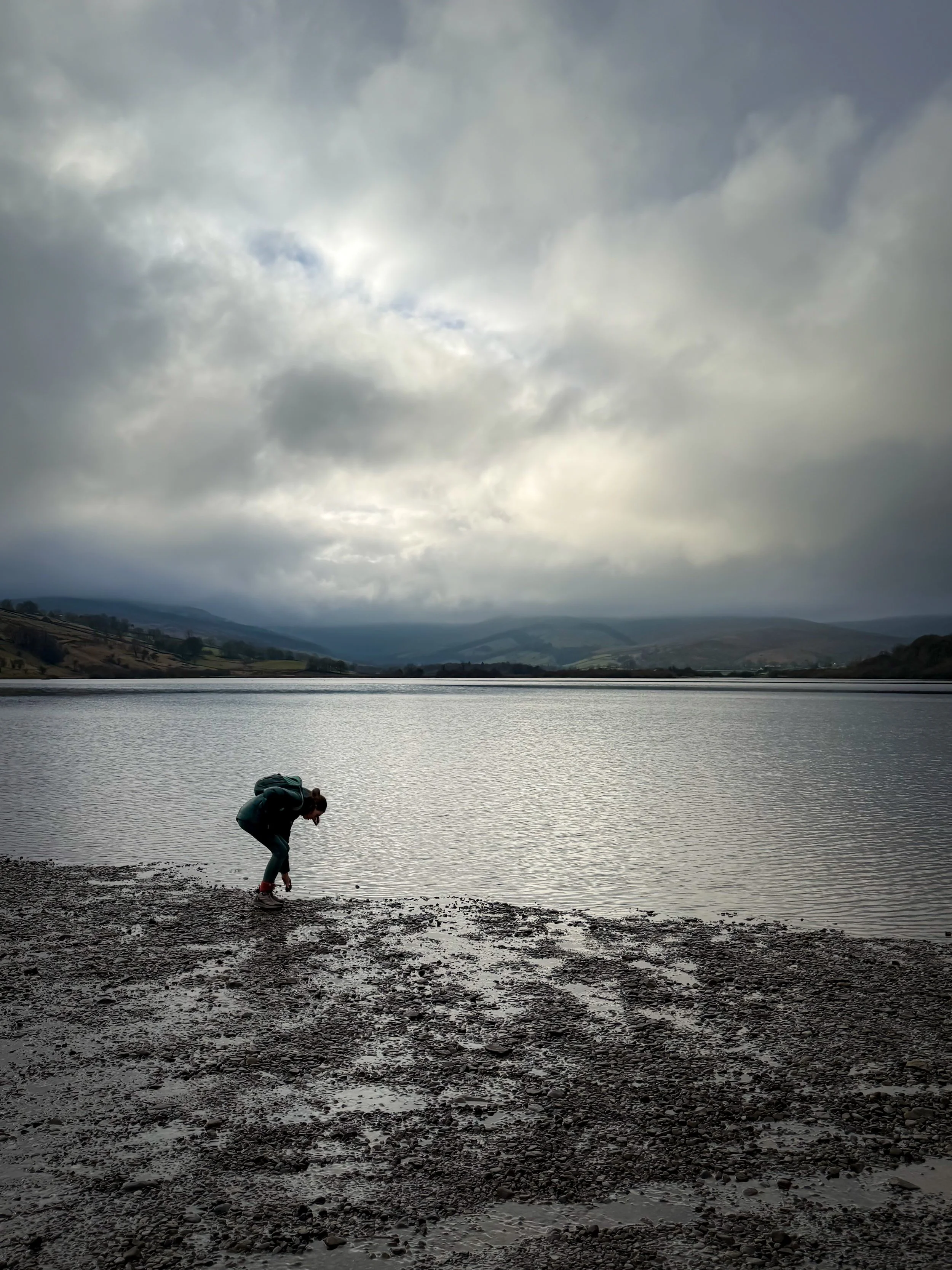 A child in silhouette bending down in front of a large still lake with hills beyon and moody grey skies with some light shining through