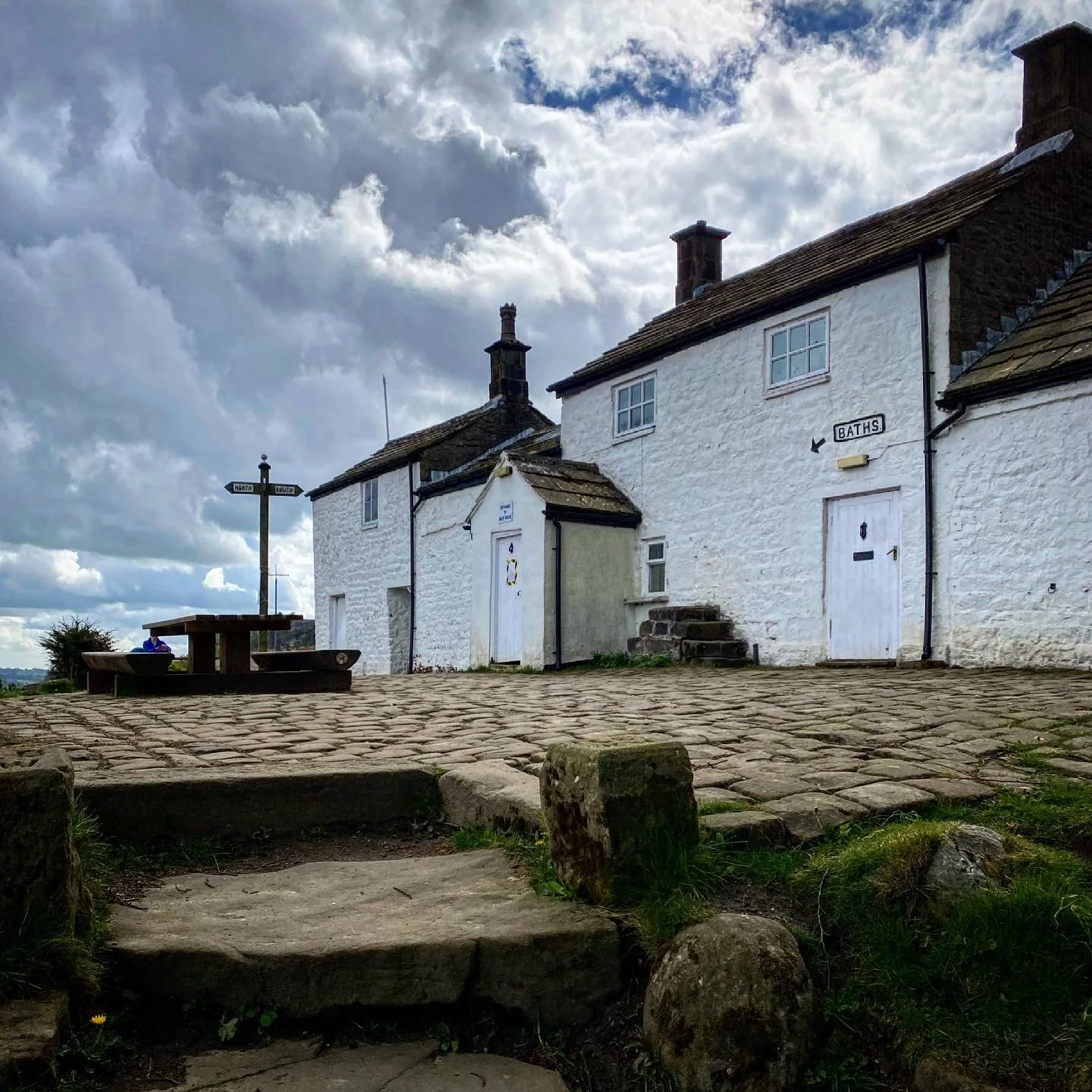 A whitewashed building with the sign baths and an arrow on it with a signpost and picnic bench with a cobbled path and under cloudy skies