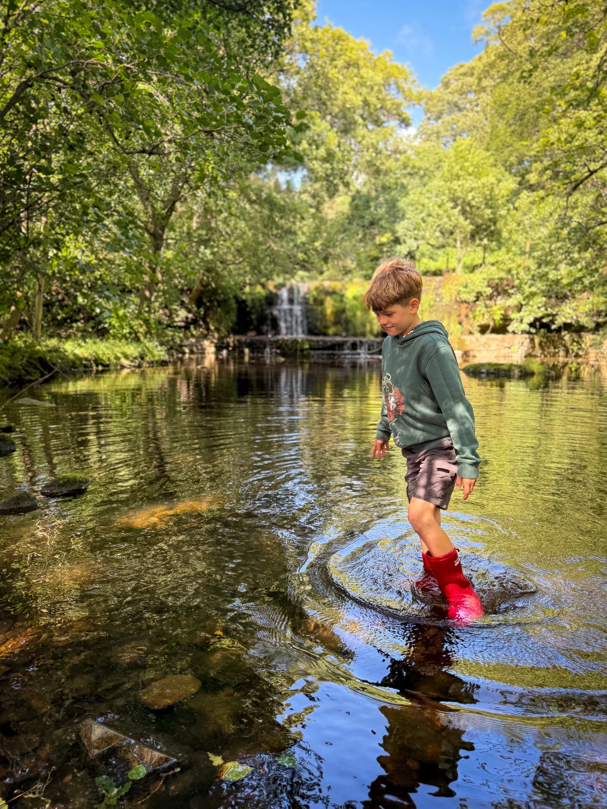 Child paddling in the water of a river in summer surrounded by trees and shrubs wearing red wellies with waterfall behind