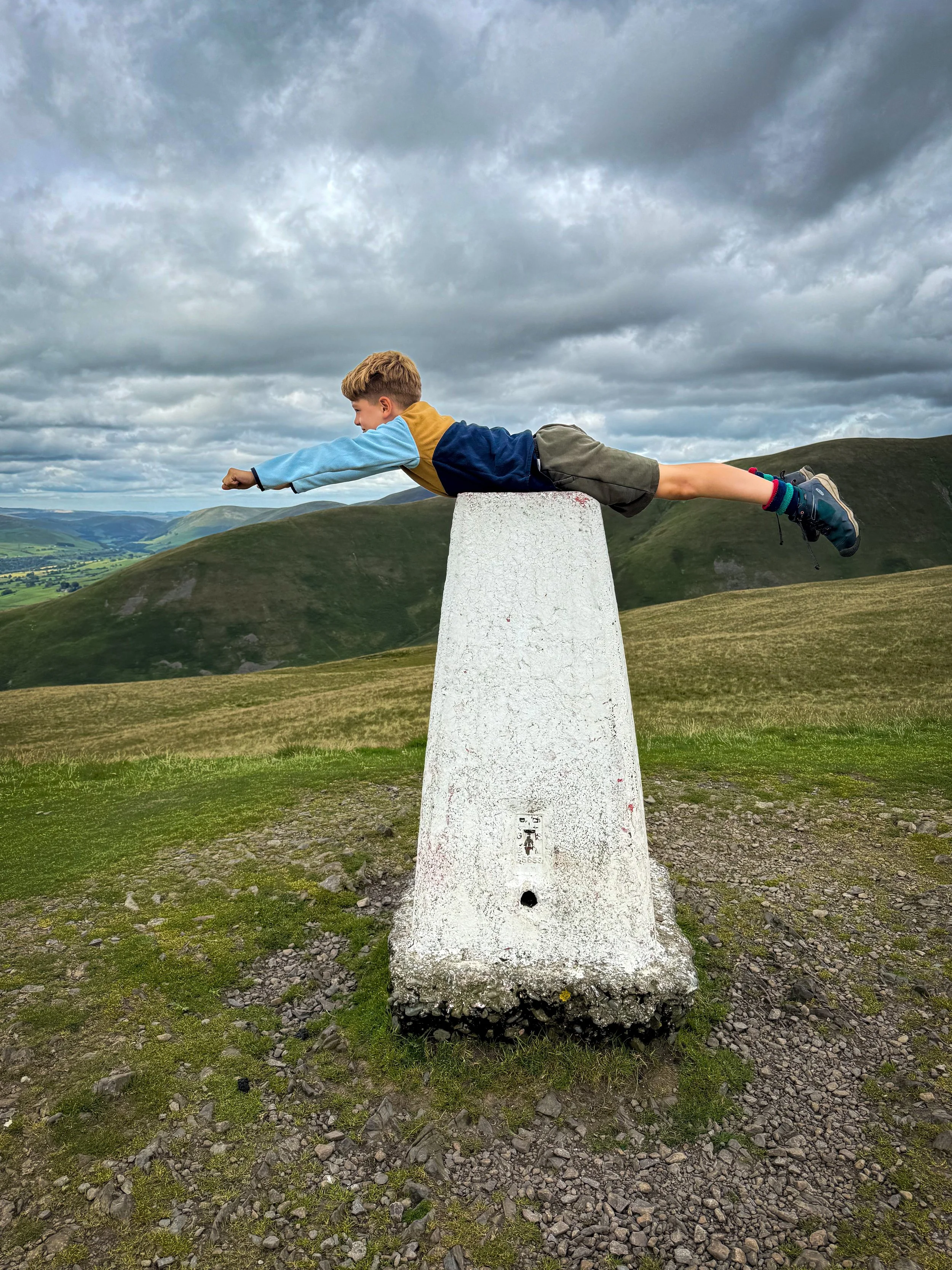 Child lying across a trig point with mountains in the background under cloudy skies