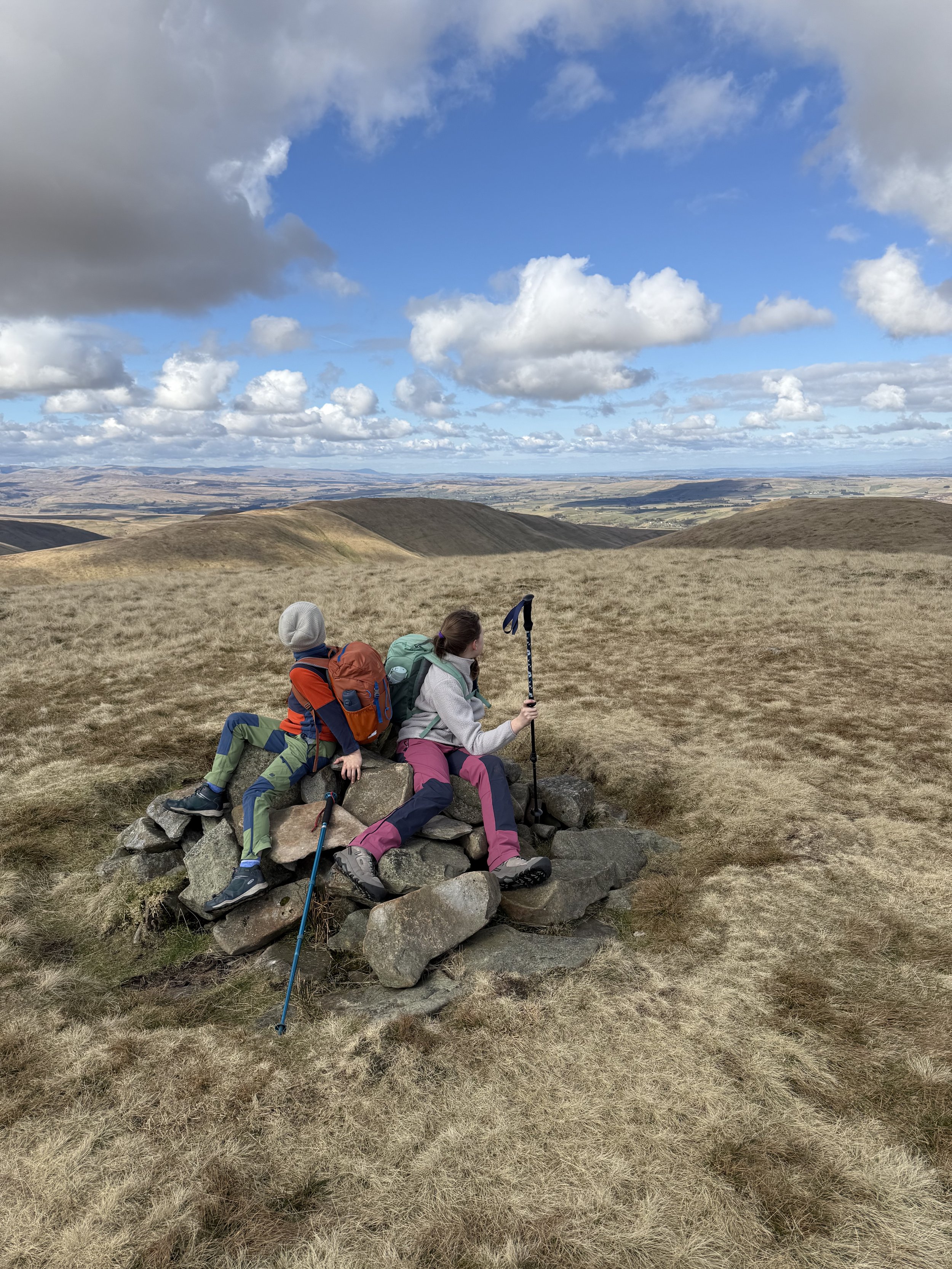 two children sitting on a summit cairn looking out at mountains in the distance