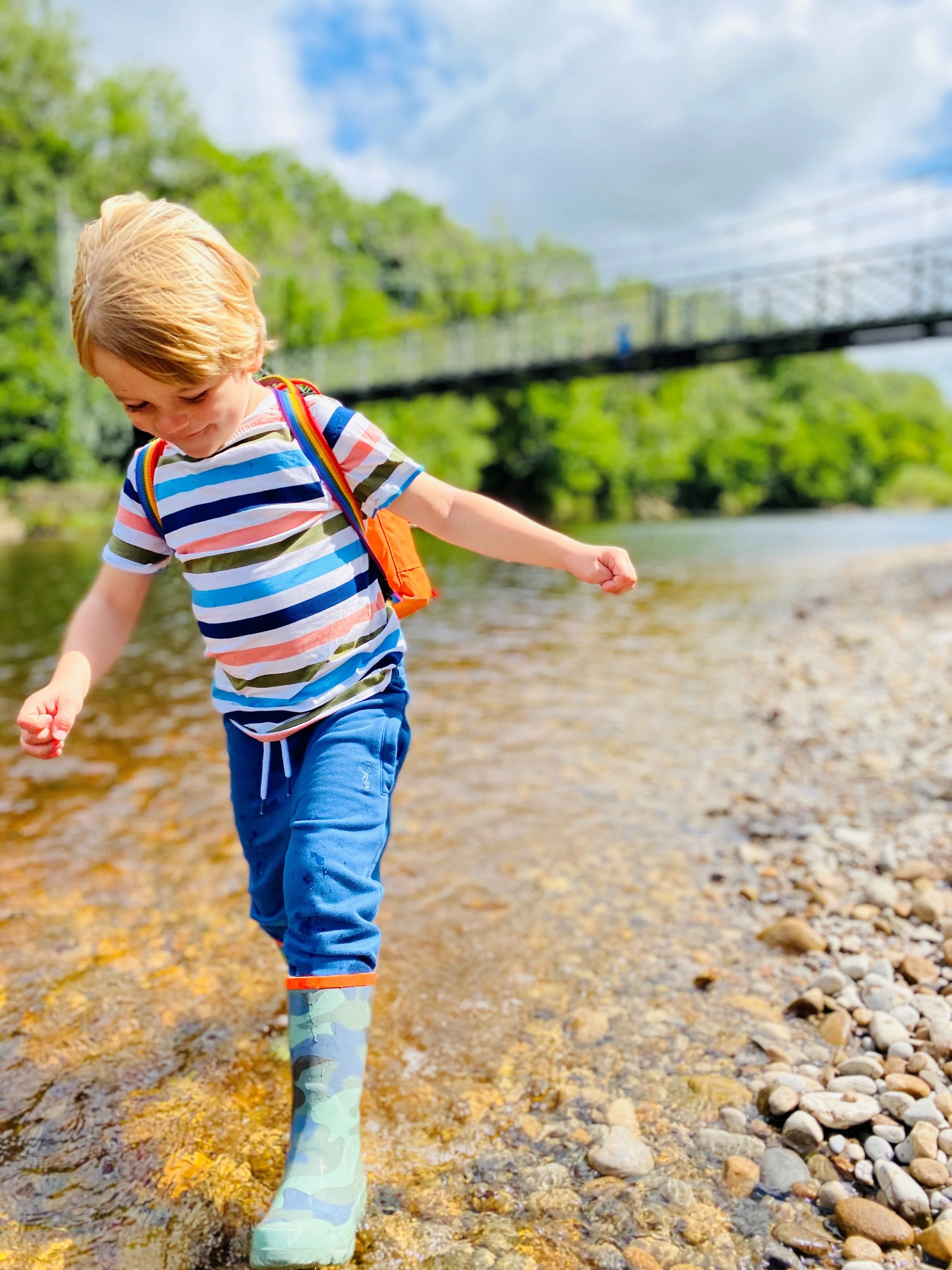 young Boy in striped tshirt and shorts paddling in wellies in the river with bridge behind