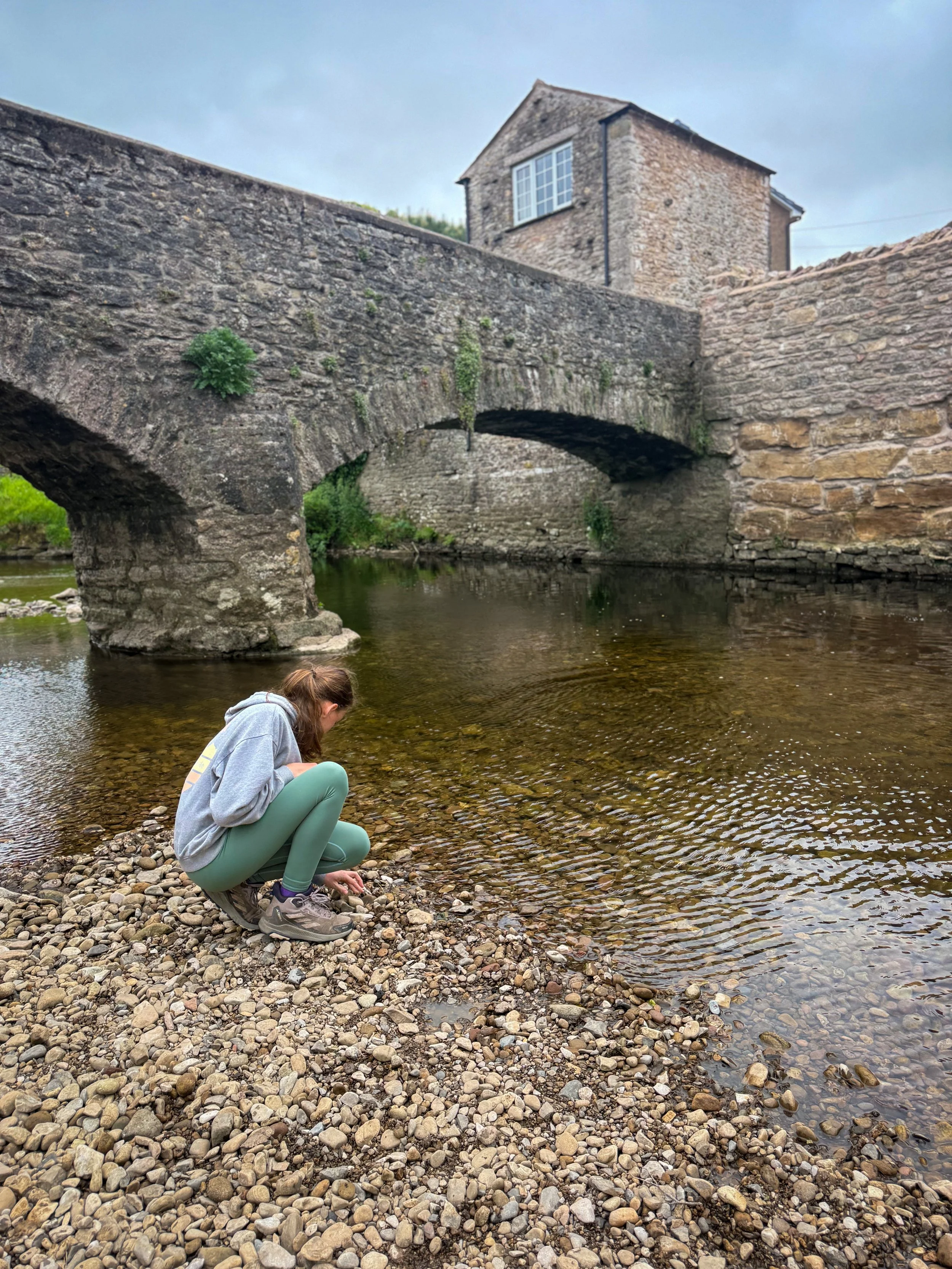 Child crouching next to the water of the river with bridge behind playing with stones