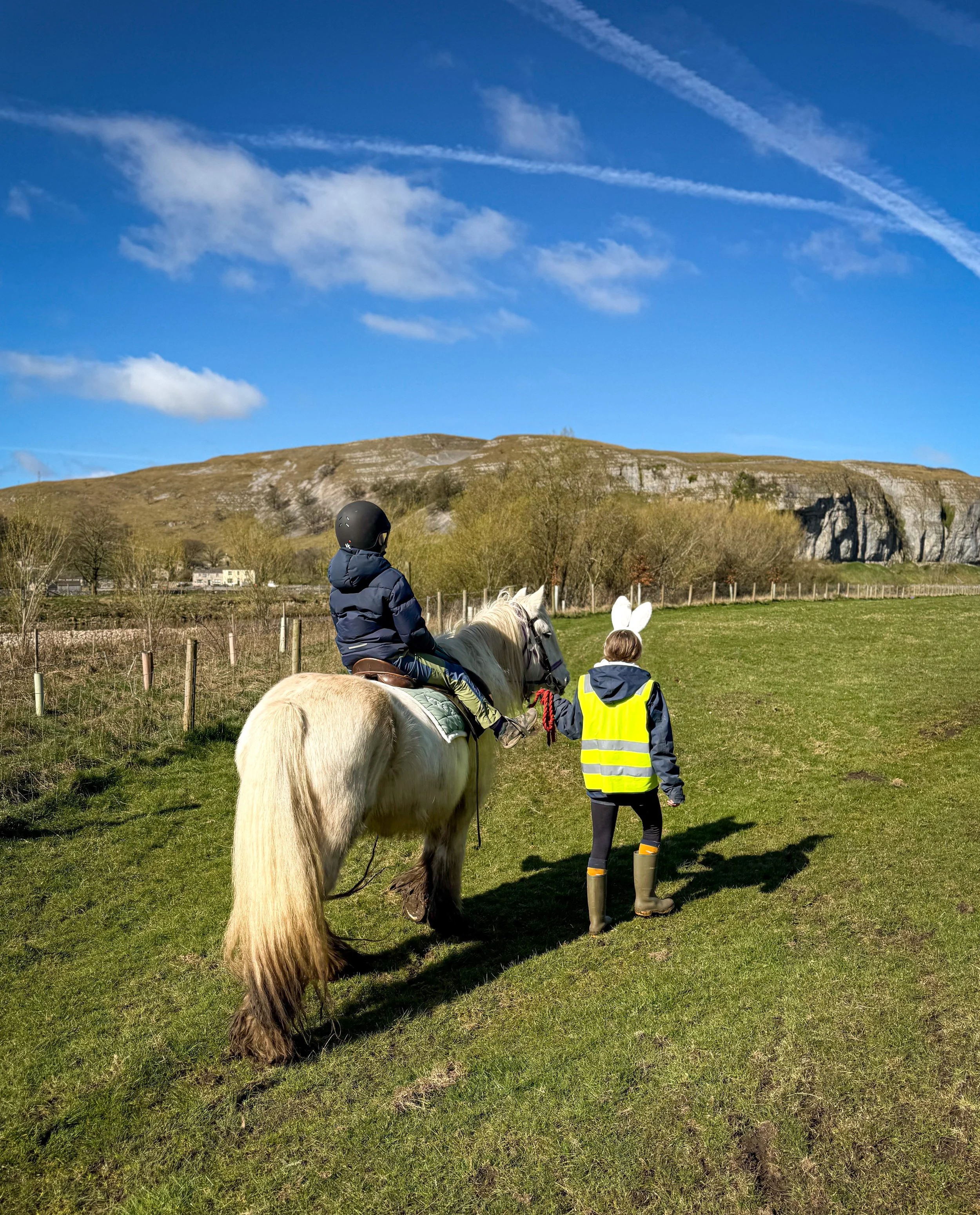 A child riding a pony being led by another child in high vis with a craggy hill beyond and blue sky