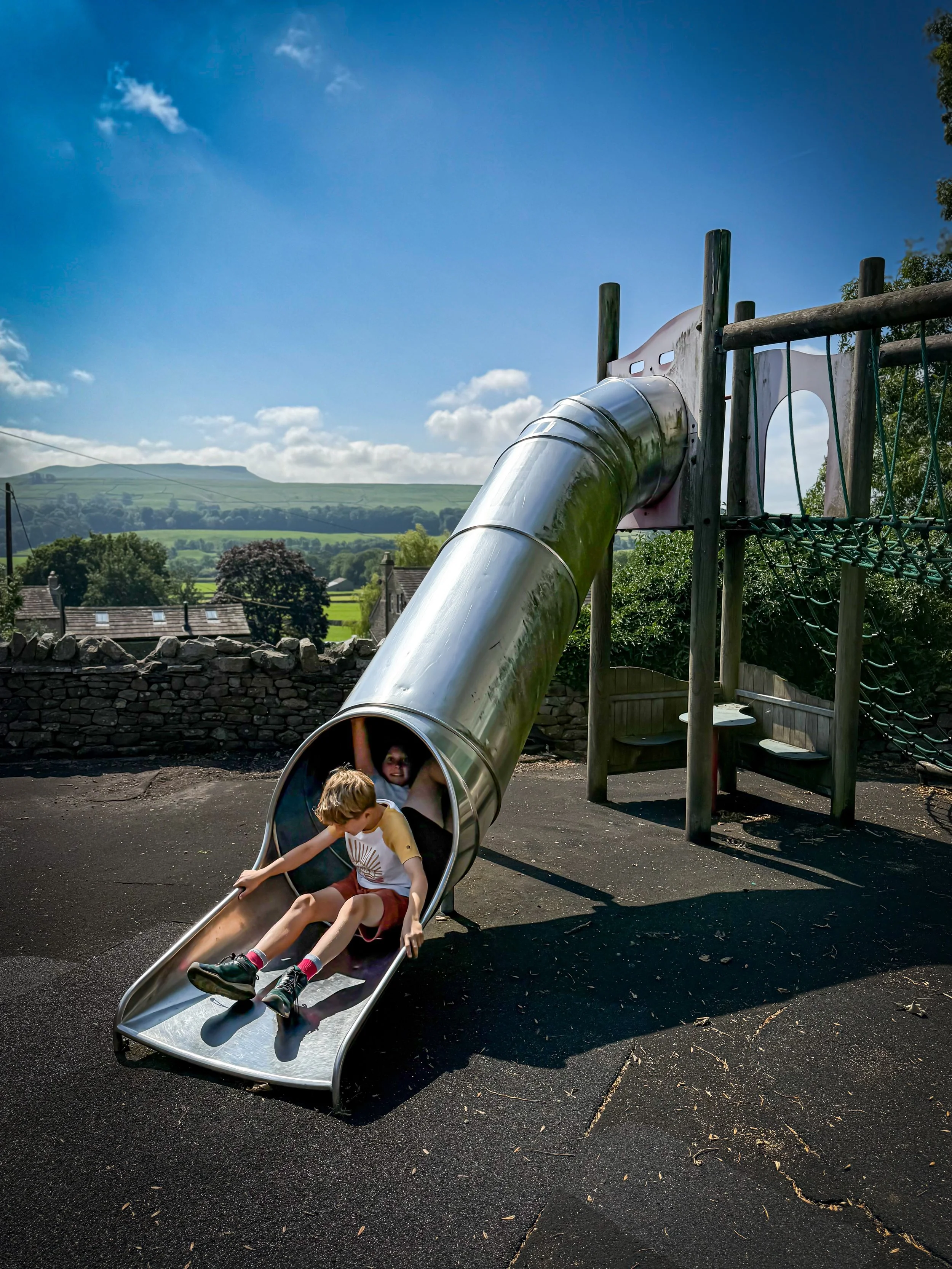 Children sliding down a tunnel slide with blue summer skies and countryside views in the distance