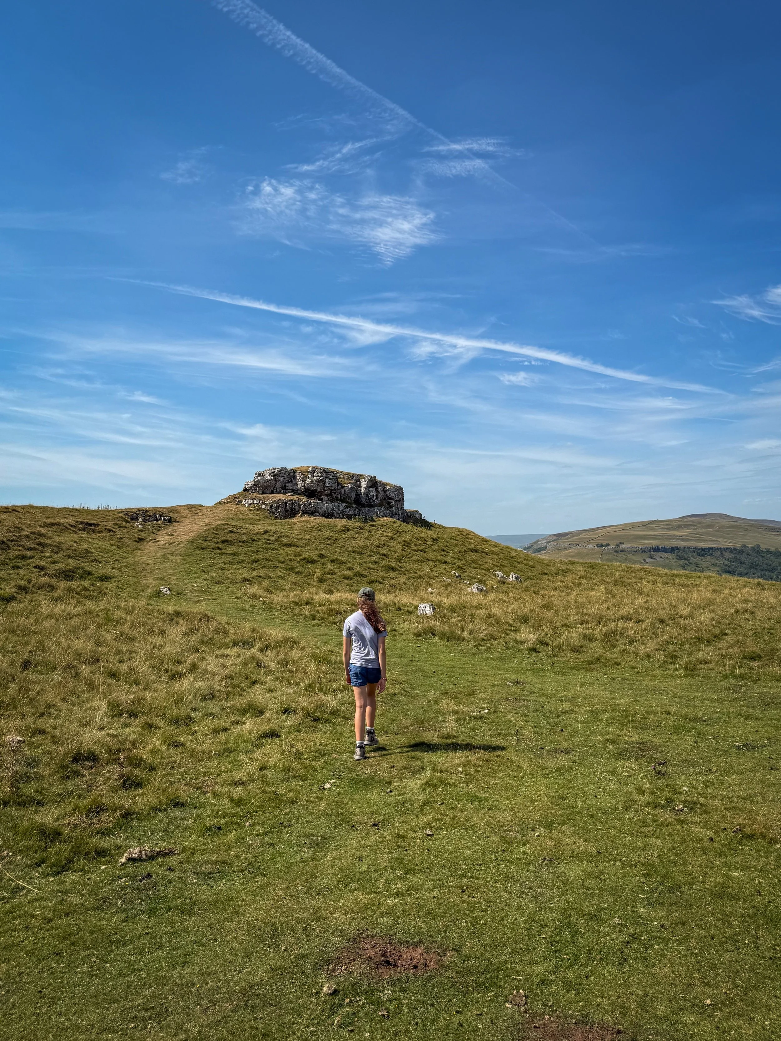 Child walking towards a rocky outcrop in the shape of a pie on top of a green grassy hill under blue skies
