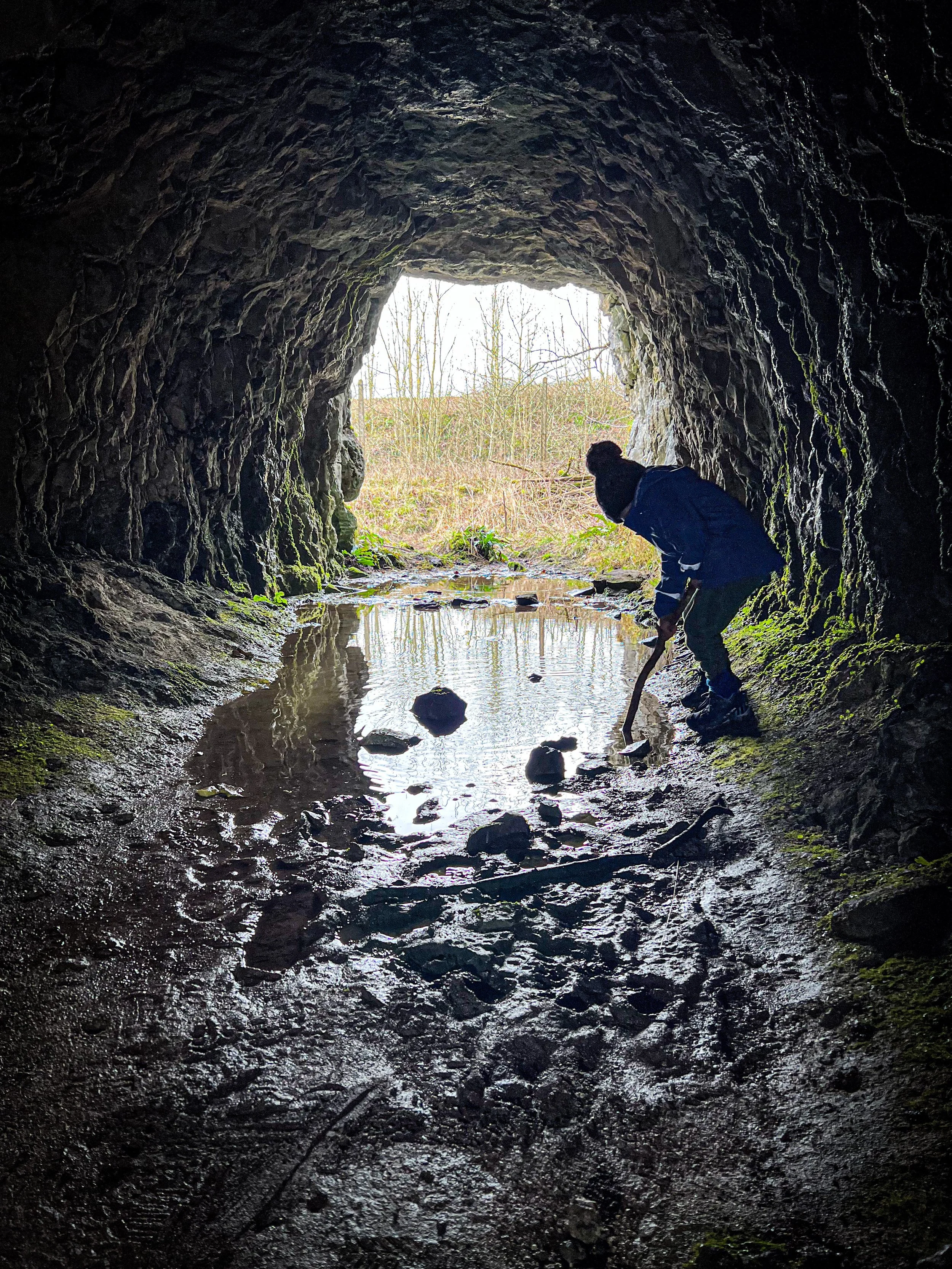 Child with a stick next to a puddle in a tunnel with light at the end.
