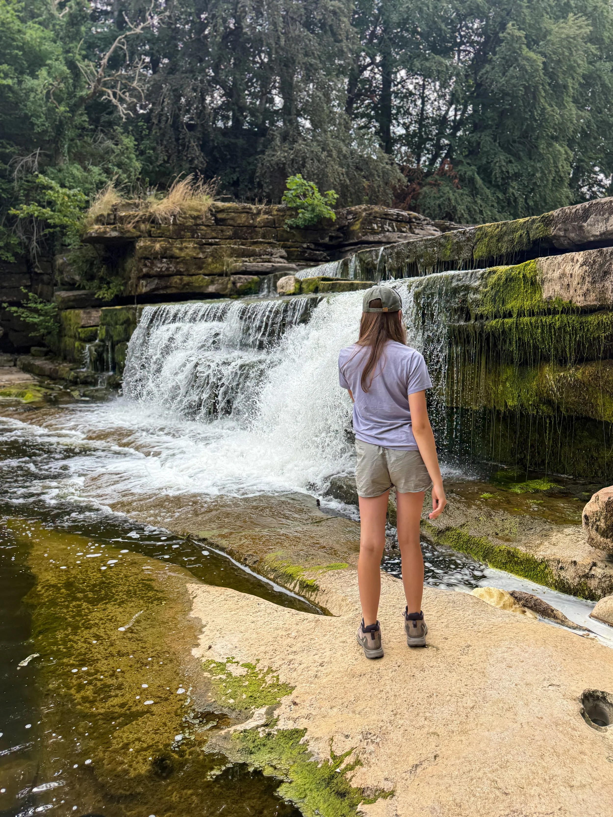 Child standing next to a waterfall in summer with trees above