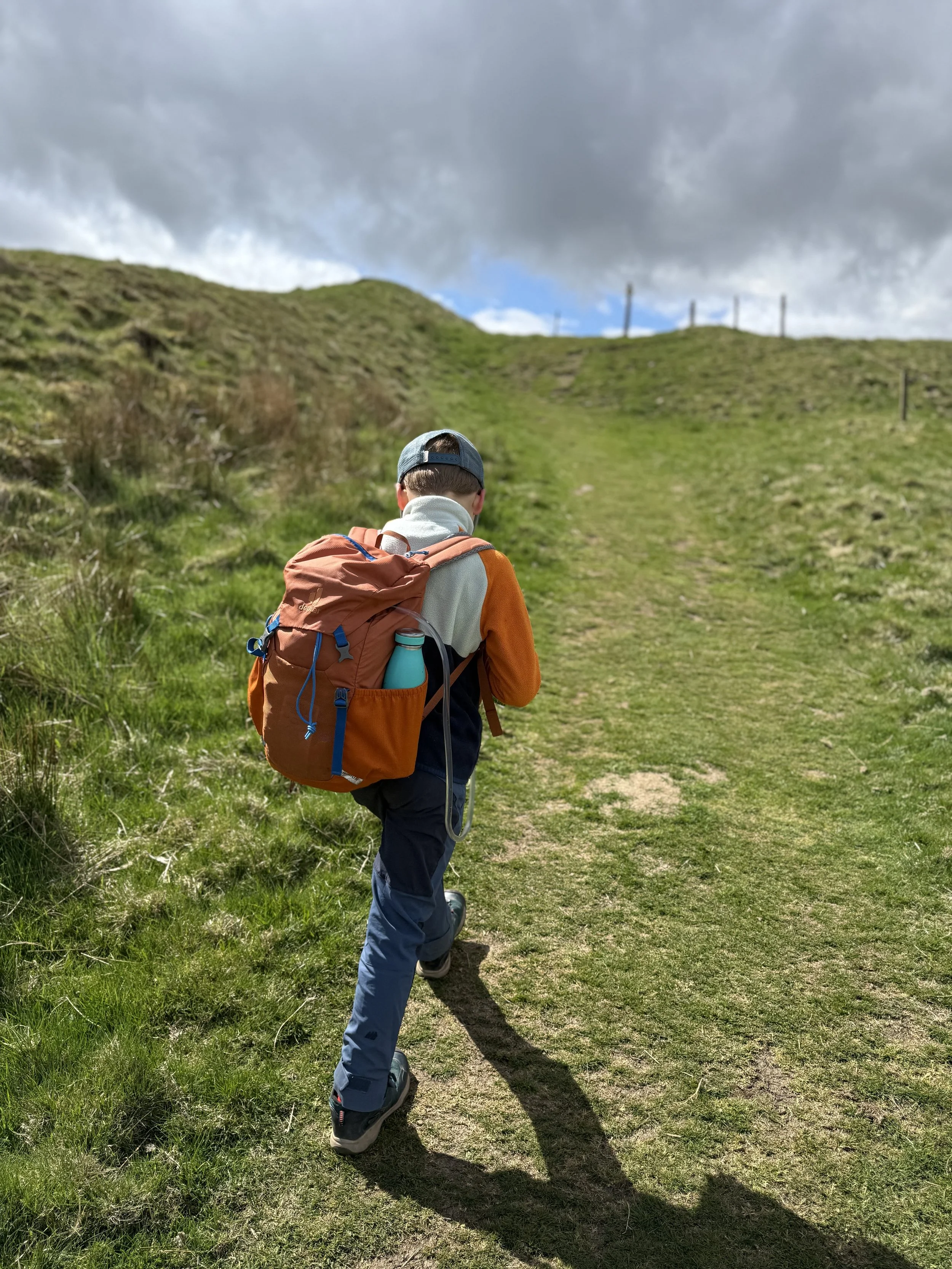 Child walking with orange backpack towards grassy hill with unusual grassy mounds beyond