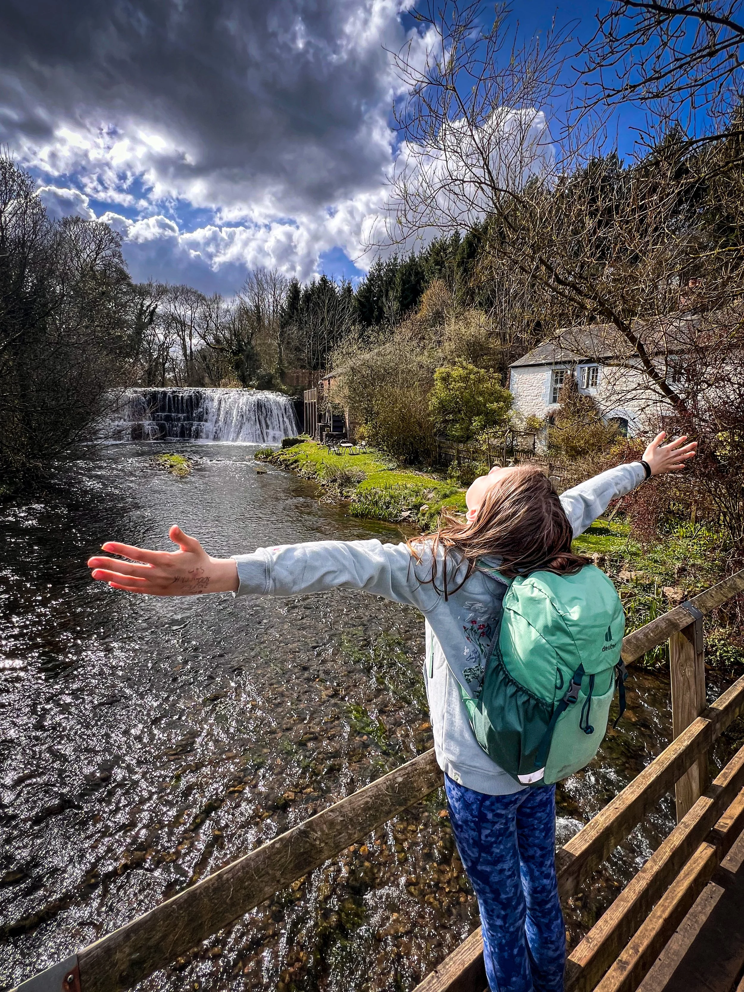 Child with arms outstretch in front of a waterfall with a cottage next door in surrounded by trees in spring