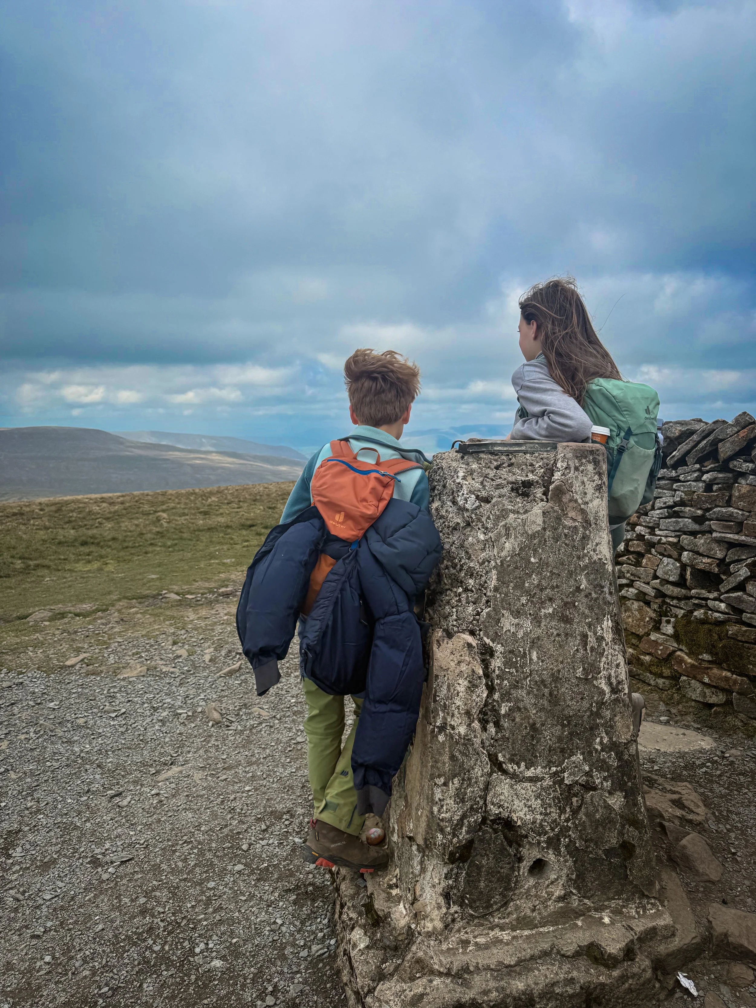 Two children standing at the summit trig of a mountain with mountain views in the distance