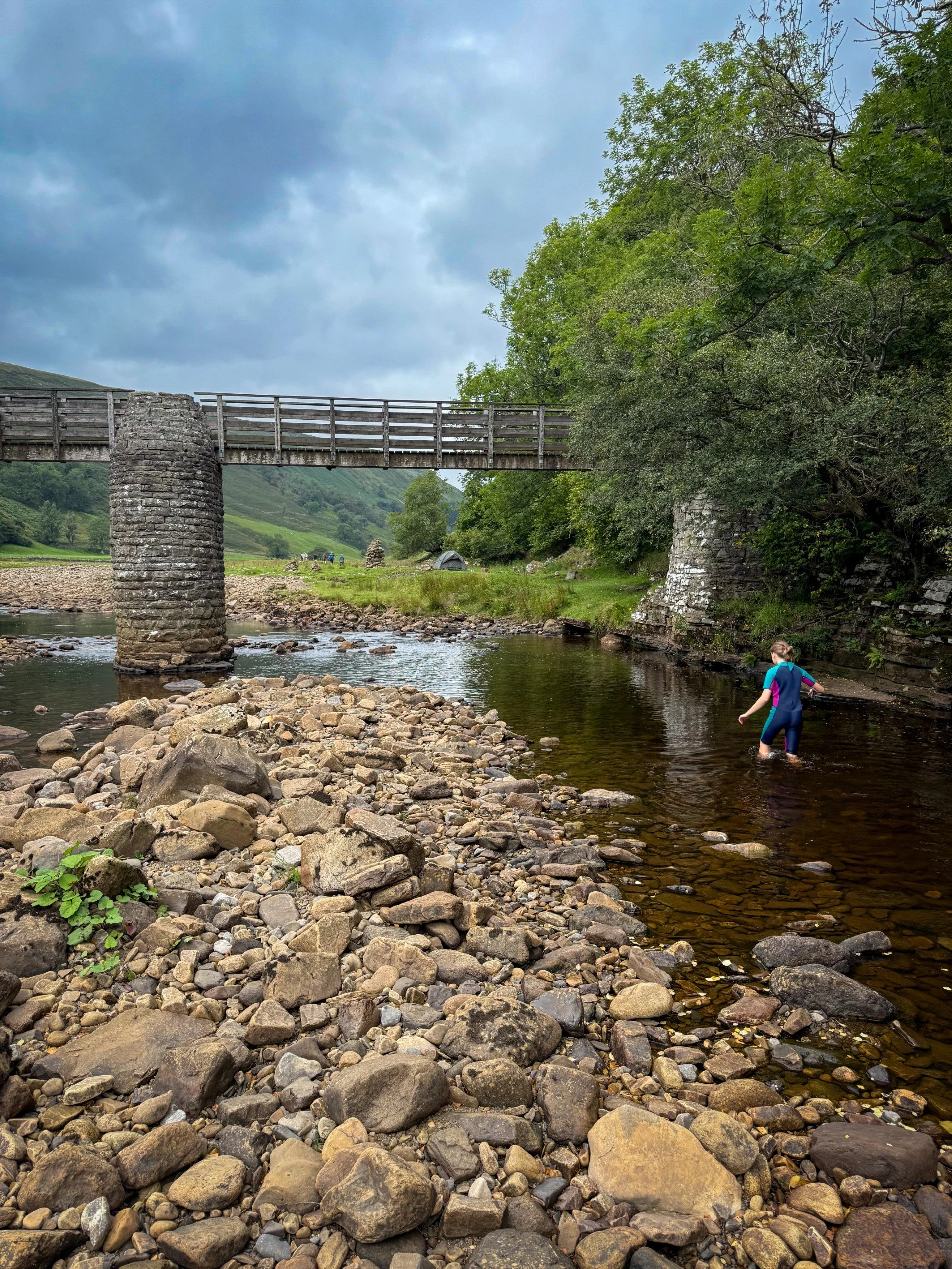 Child wading through a river with a bridge behind made of wood and dry stone pillars in the summer with trees and a grassy hillside behind