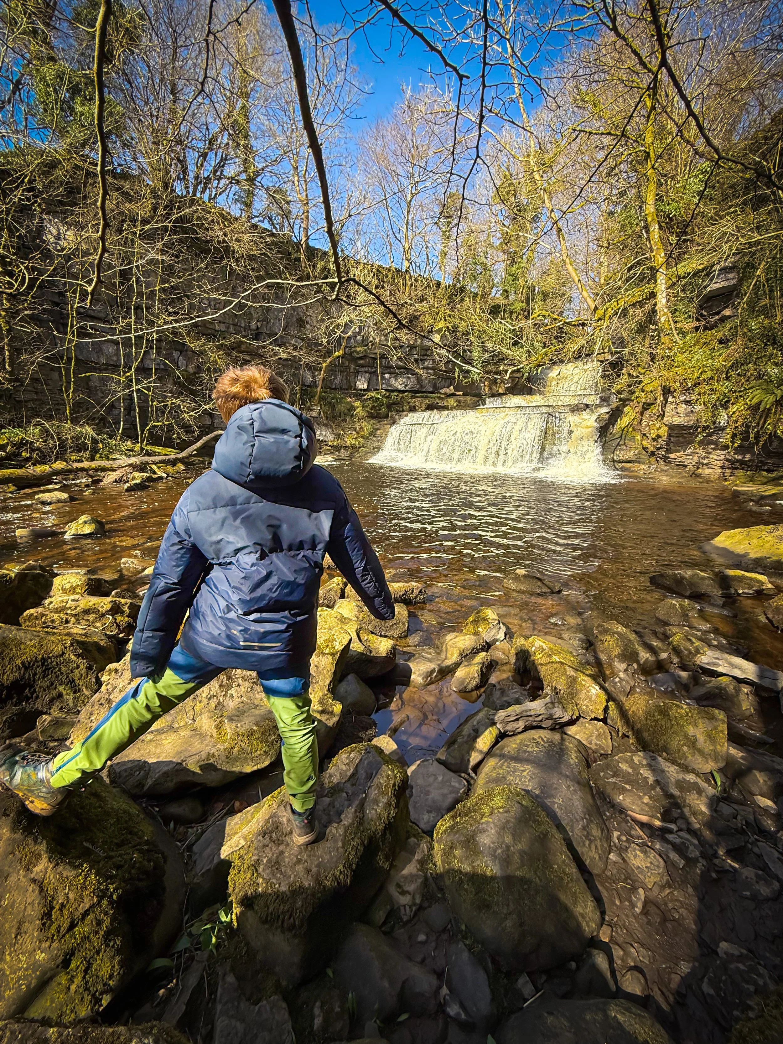 Child standing next to a waterfall in spring surrounded by trees and rocks