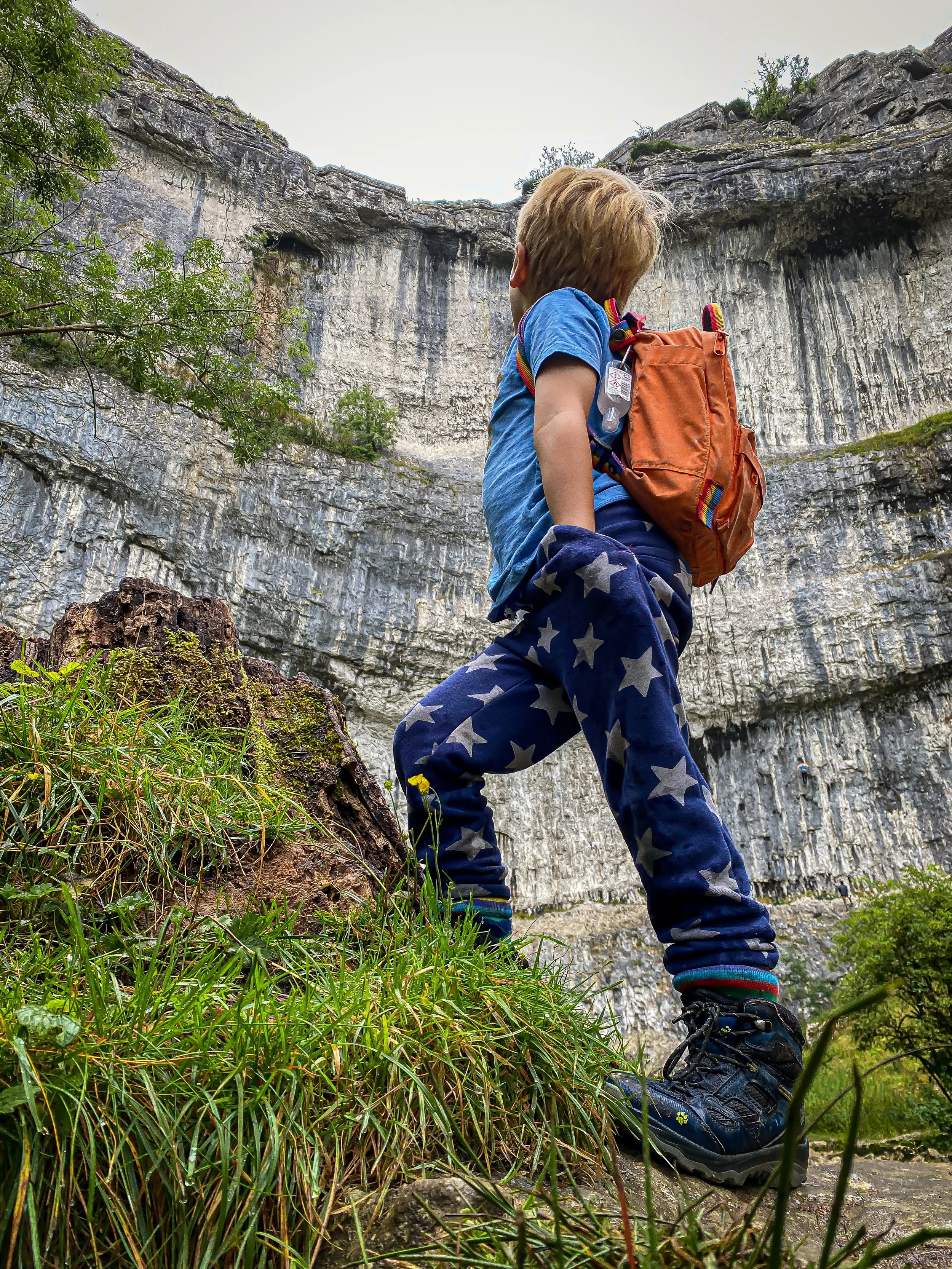 Child wearing star print joggers and a blue tshirt with an orange backpack looking up at a large white rocky cliff face with green grass in the foreground