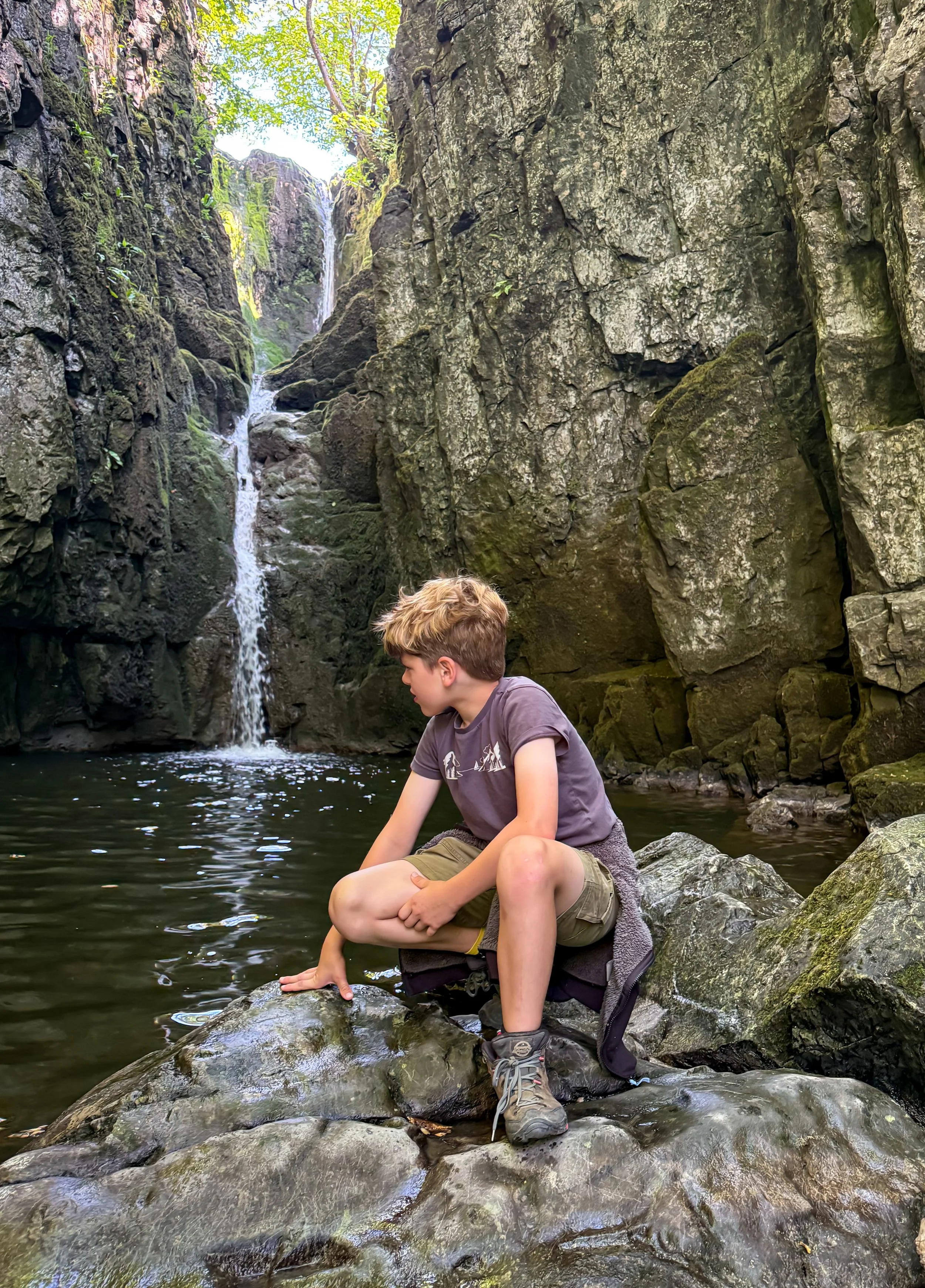 Child sat on a rock next to a waterfall in a rocky gorge in summer