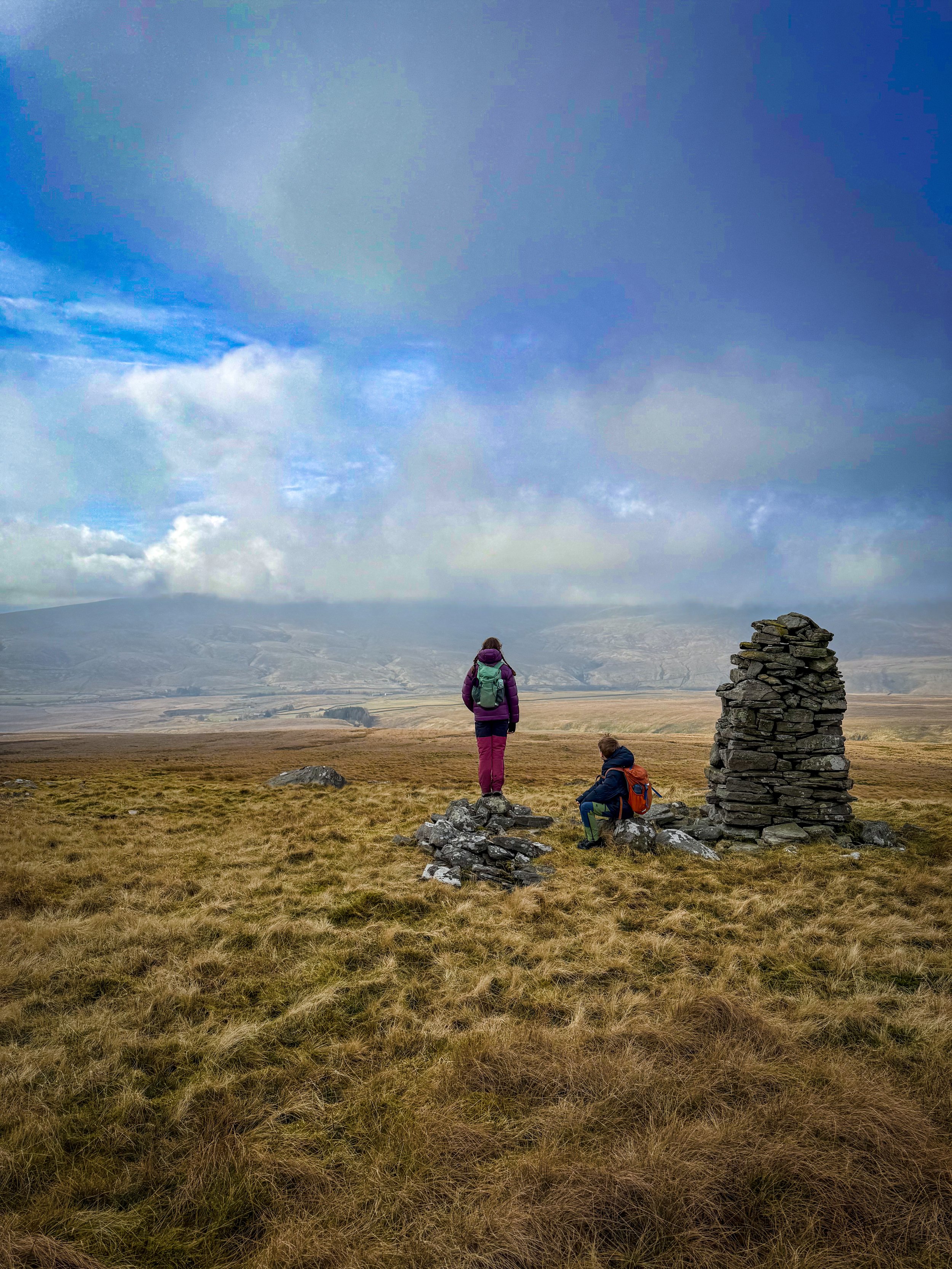 a child sat and one child stood next to a large stone built cairn looking down from the top of a mountain to the valley and mountains beyond