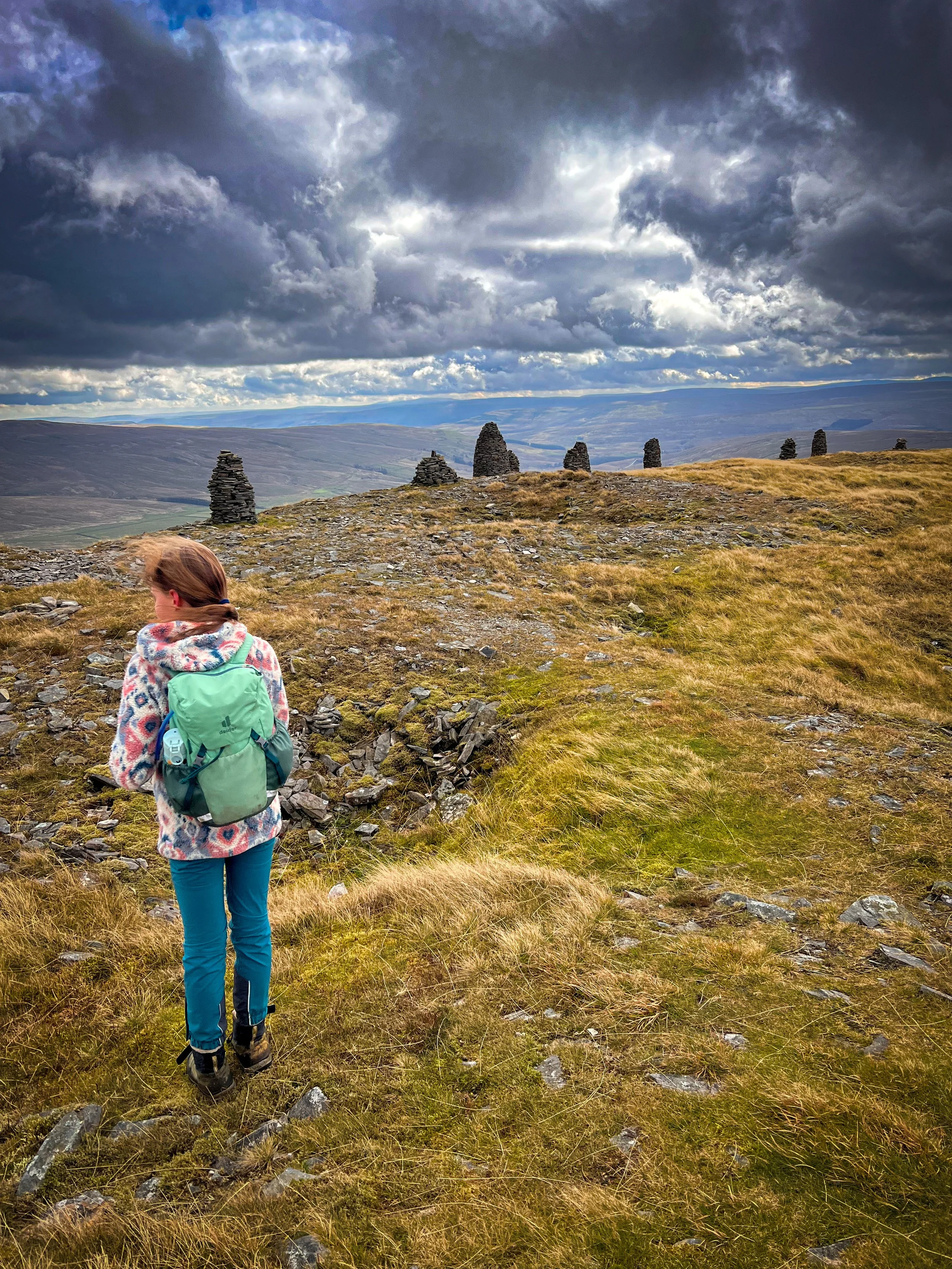 Child standing in a fleece and walking trousers with cairns behind with cloudy skies and mountains