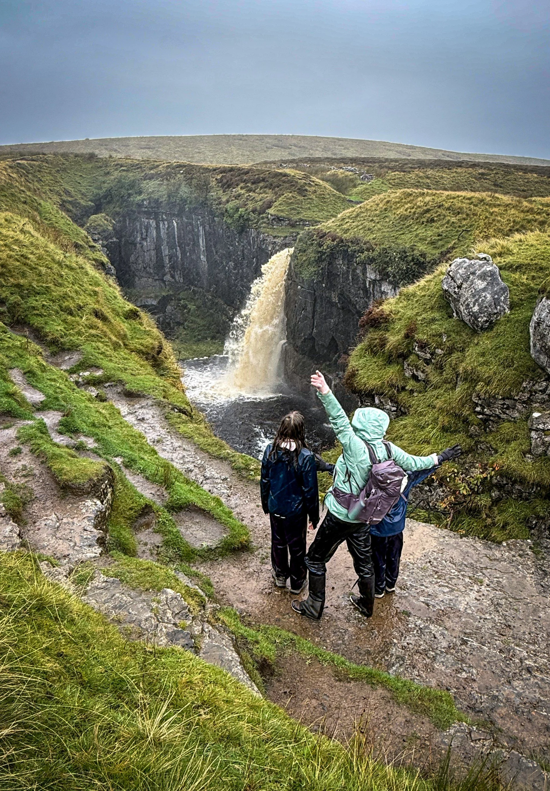 One adult and two children standing next to a huge collapsed cavern with a massive waterfall flowing into it with rain
