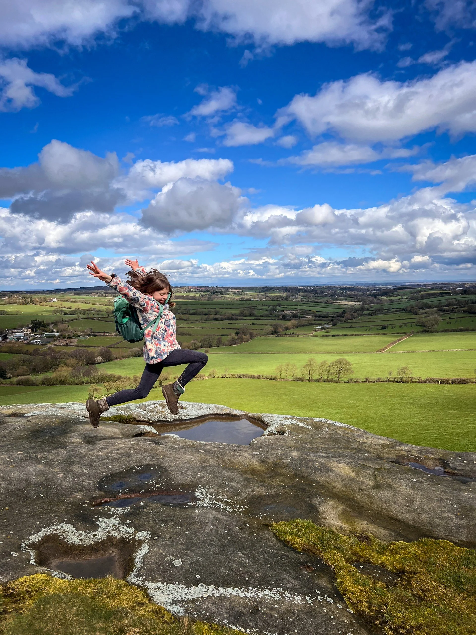 Child leaping with arms in the air on a rock with green grassy fields in the background and blue skies above with white fluffy clouds