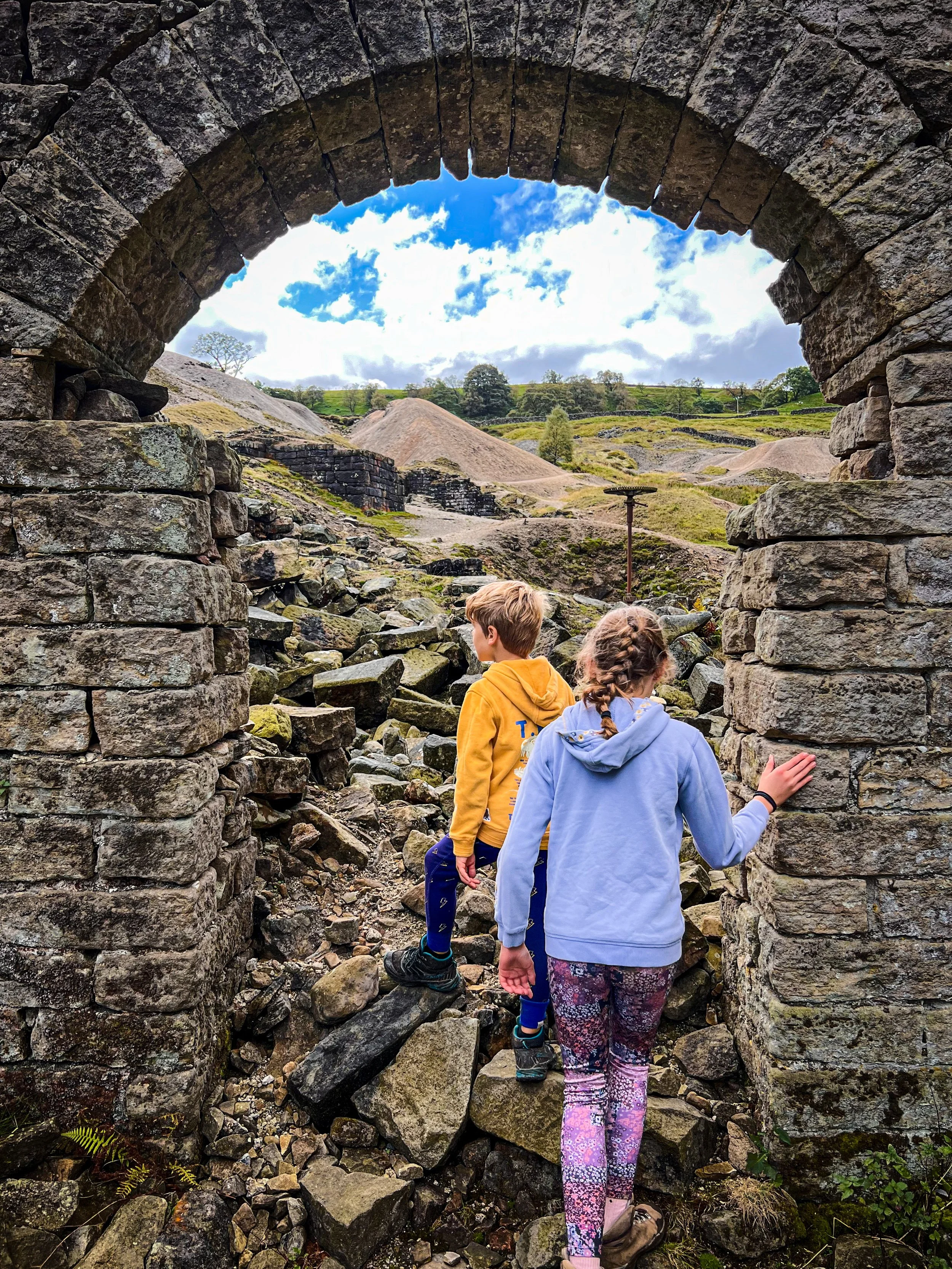 Two children walking through a dry stone archway with ruined building behind