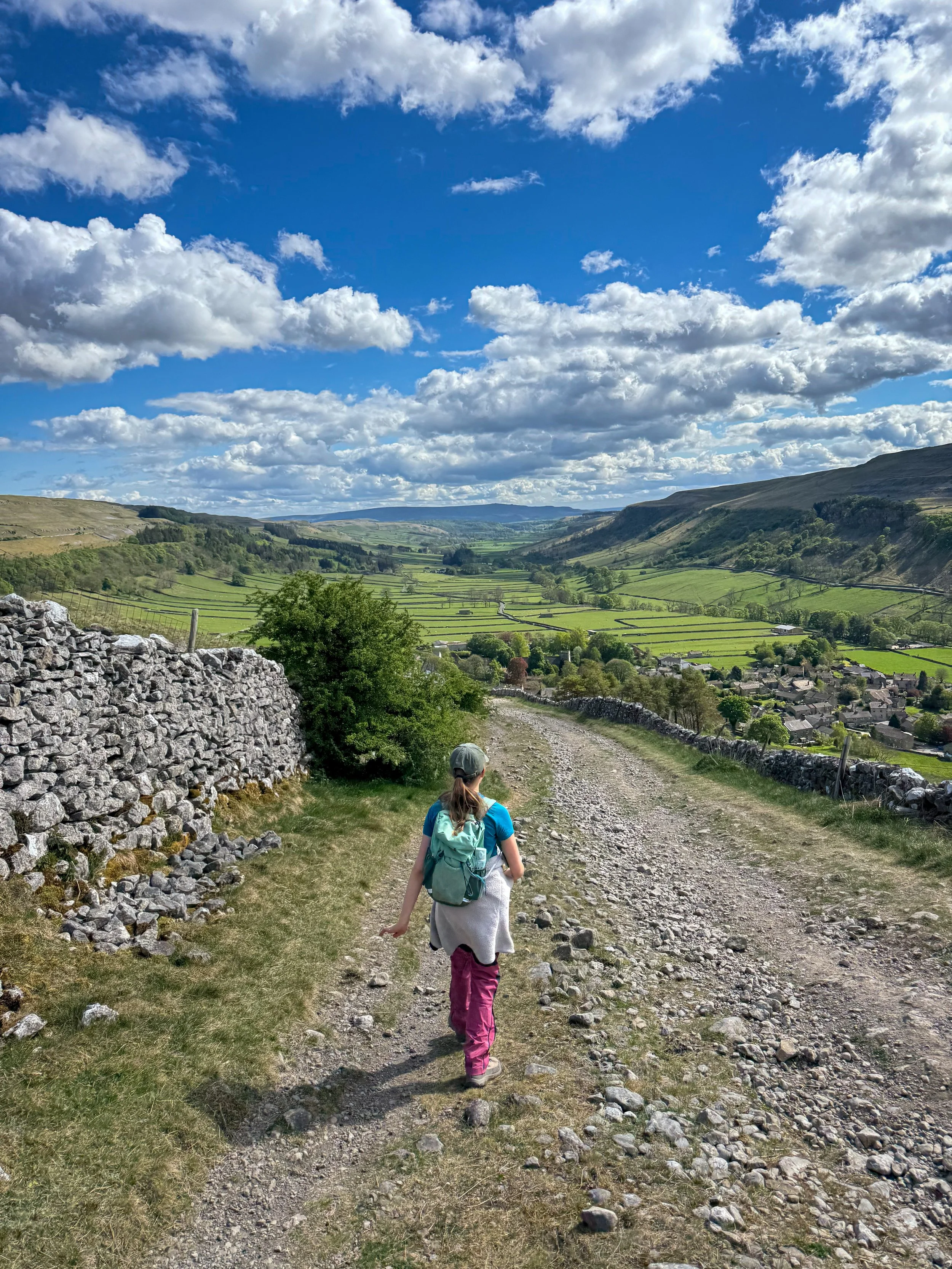 a child wearing pink trousers walking towards a village on a wide walled track under blue skies in summer