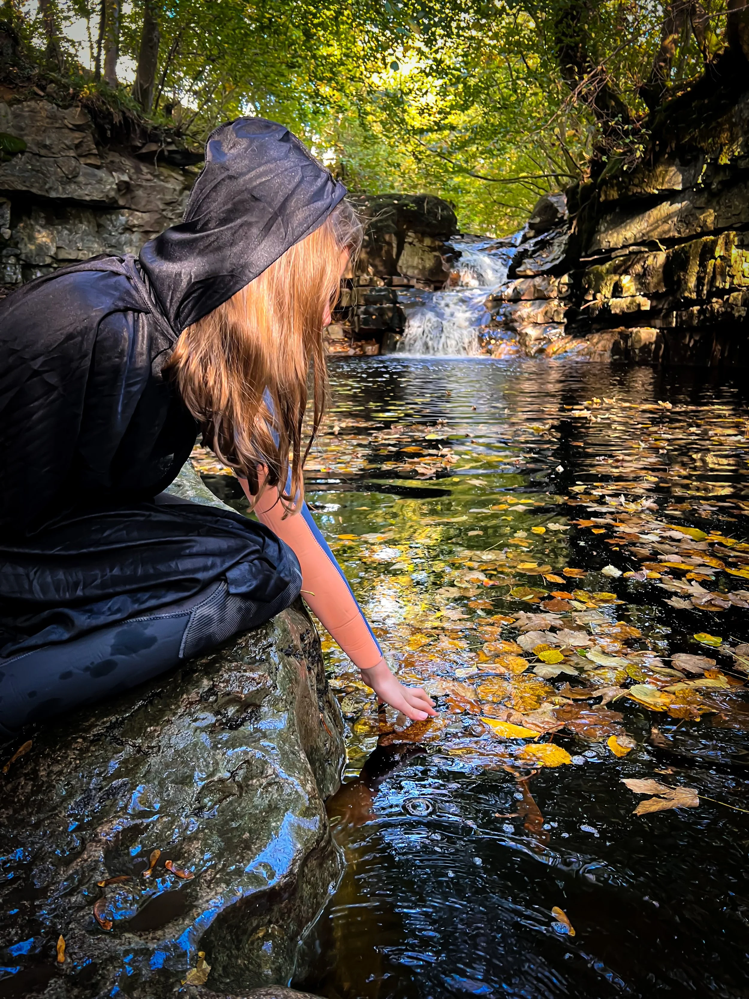 Child wearing a wetsuit and witch's cloak with her hand in water filled with autumn leaves with a waterfall behind