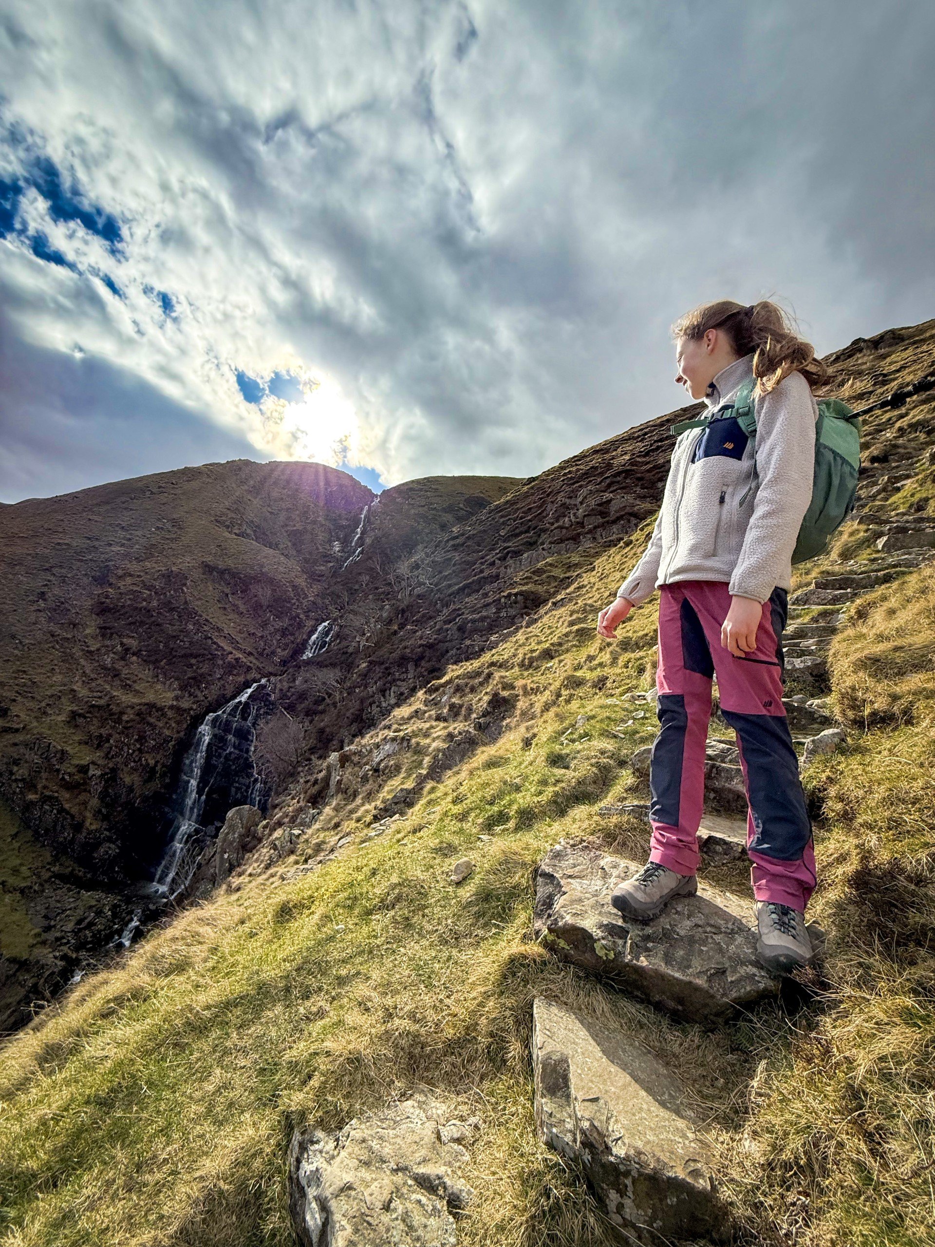 Child standing in front of a waterfall  with sun peeping on fellside