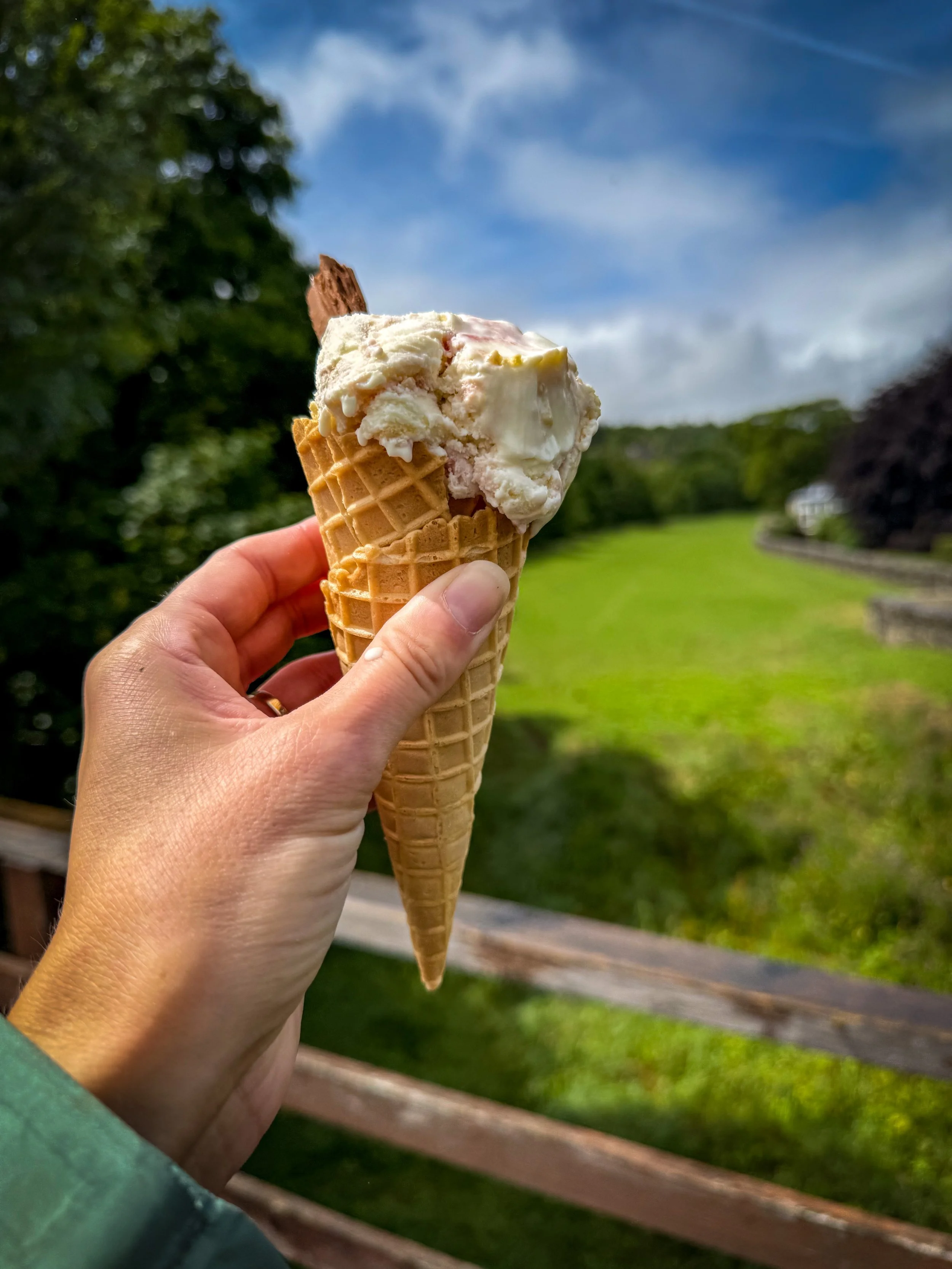 Hand holding ice cream with a waffle cone and chocolate flake in the top over a bridge with sunshine