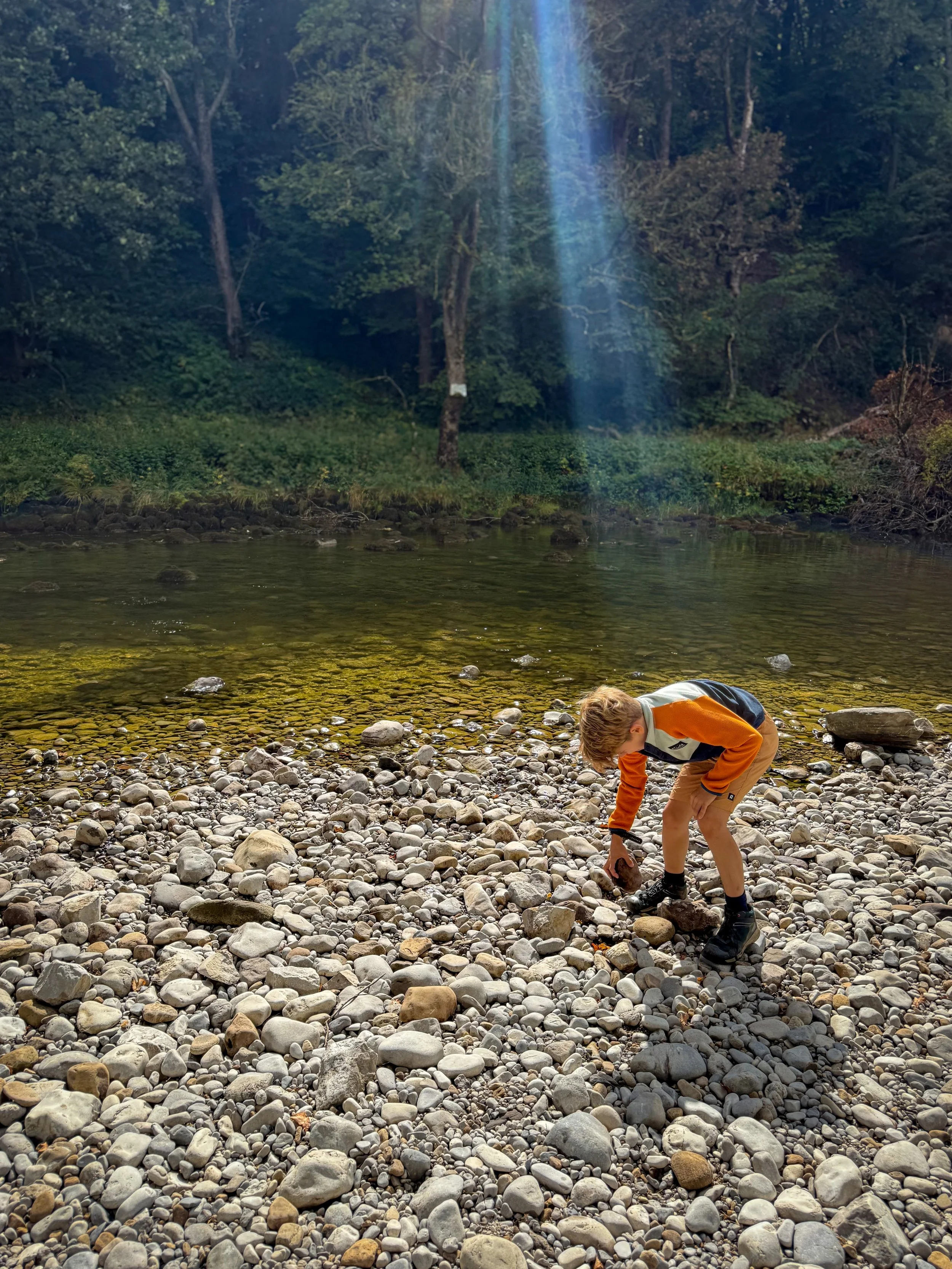 Child playing by the water of a river with thick trees the other side and rocks in the foreground
