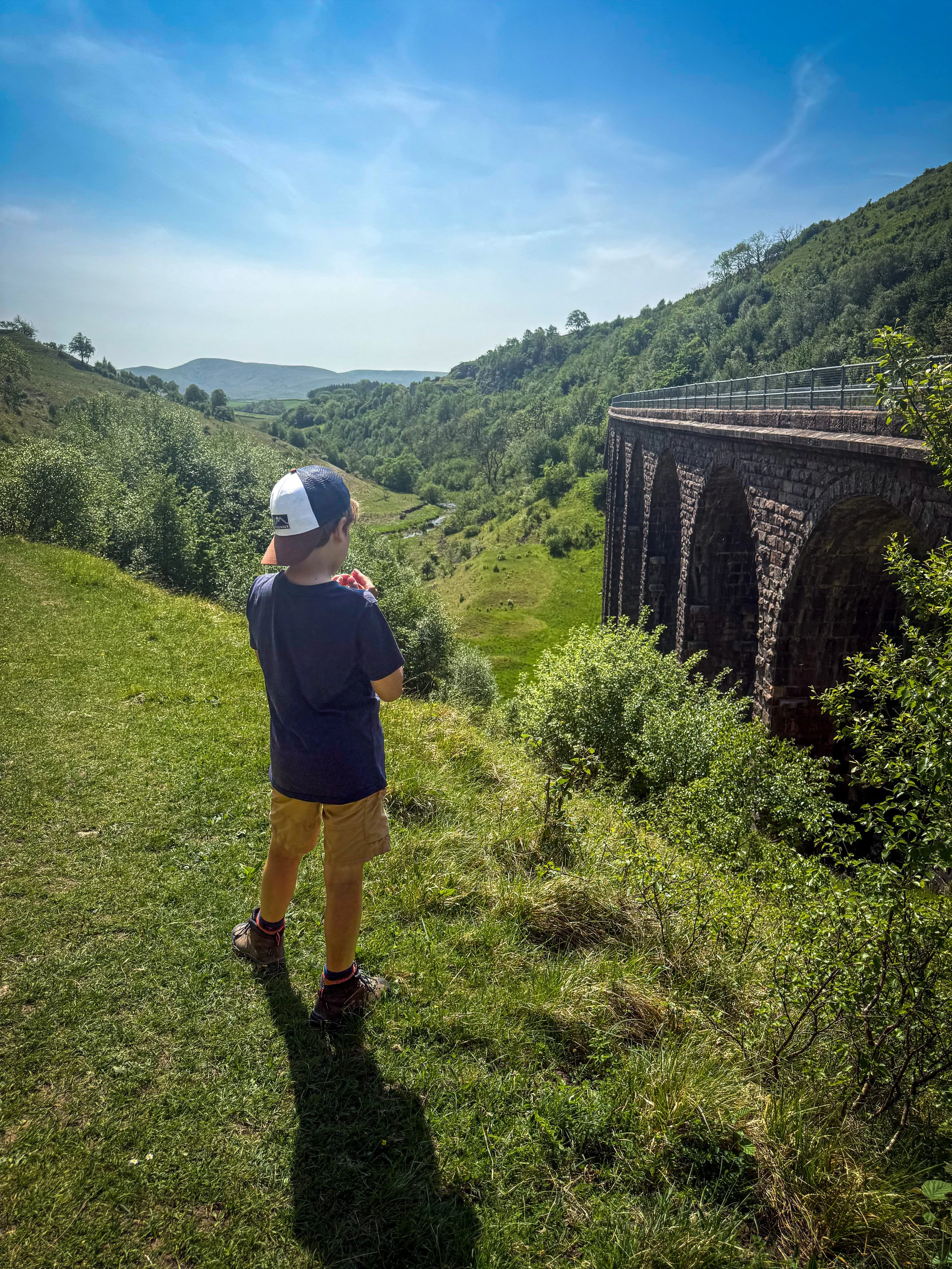child in backwards cap standing on a grassy bank high up next to a viaduct with mountain views beyond